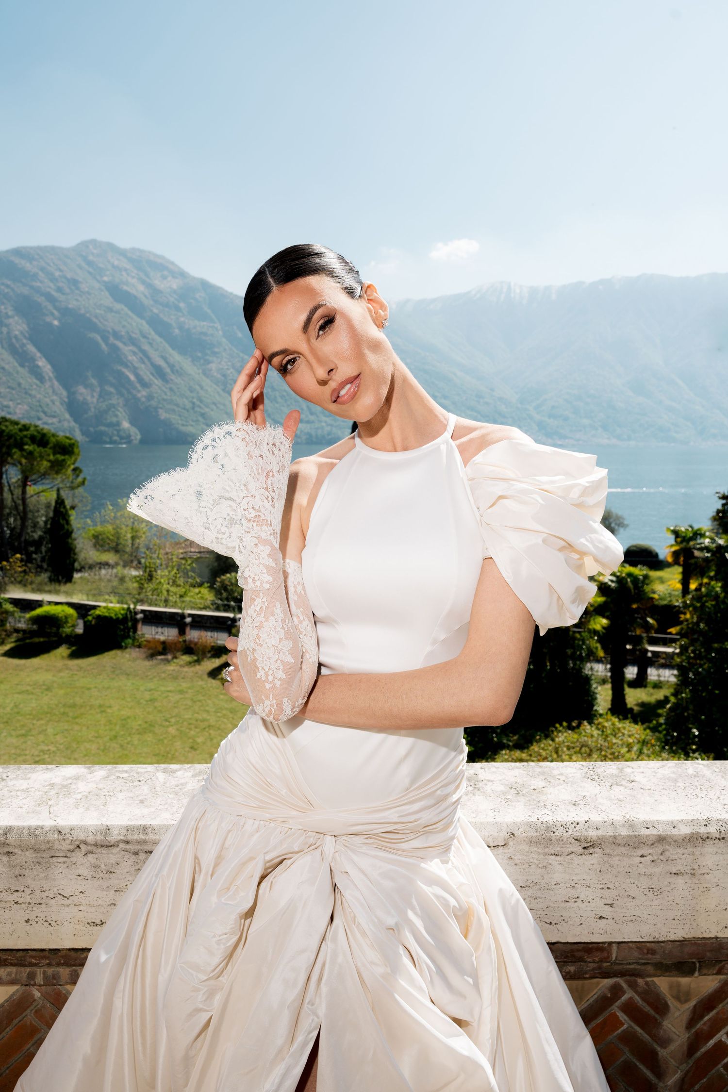 Bride in elegant white gown with lace gloves poses on balcony with mountain lake backdrop.