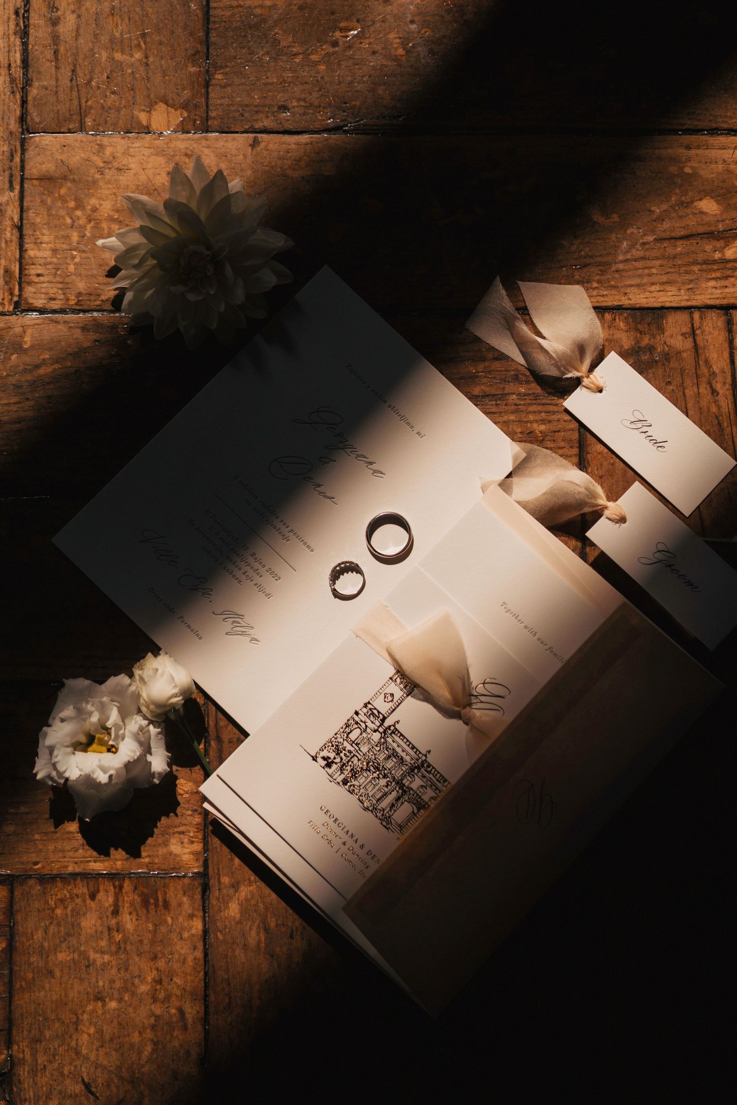 Wedding rings cast dramatic shadows on white paper invitations against rustic wooden table surface in moody lighting.