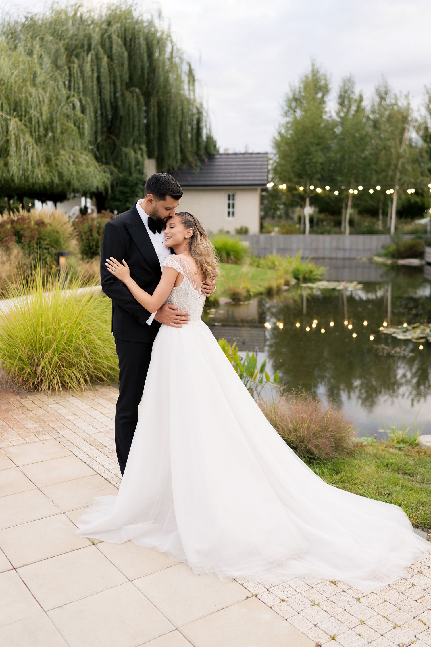 A romantic wedding couple embraces by a pond with weeping willows and string lights creating an enchanting atmosphere.