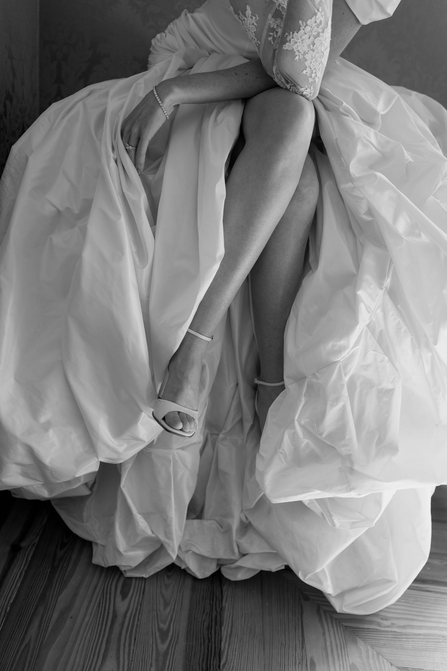 Black and white photo of a bride in a flowing white wedding gown seated on wooden floor, legs visible beneath voluminous skirt.