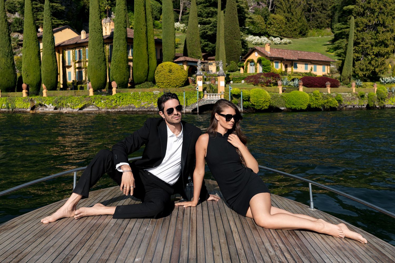 Elegant couple in black outfits sitting on a boat dock with a luxury lakeside villa and cypress trees in the background.