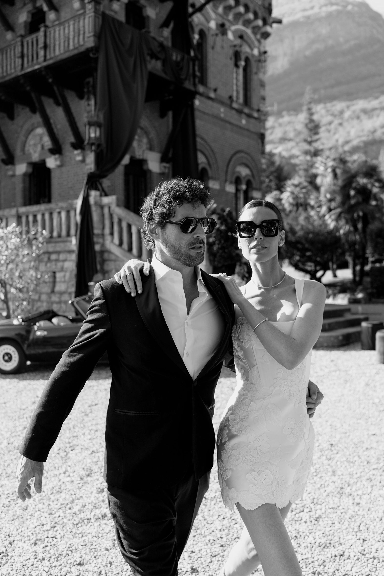 Black and white photo of stylish couple in sunglasses, bride in white dress and groom in suit, walking before elegant villa.