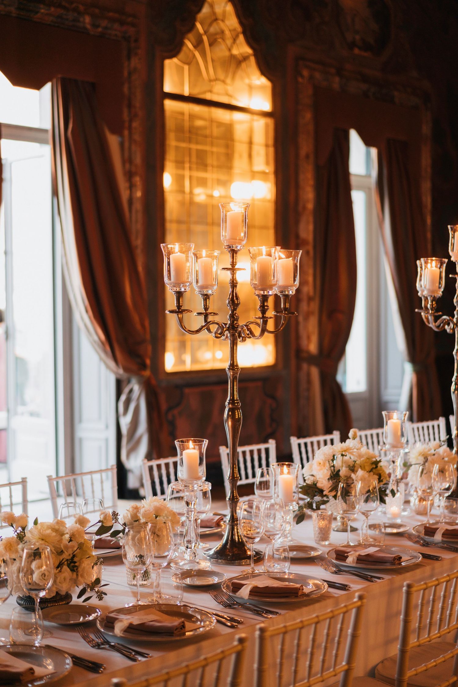 Elegant wedding reception table with tall candelabra centerpieces, brown drapes and golden lighting in a luxury venue.