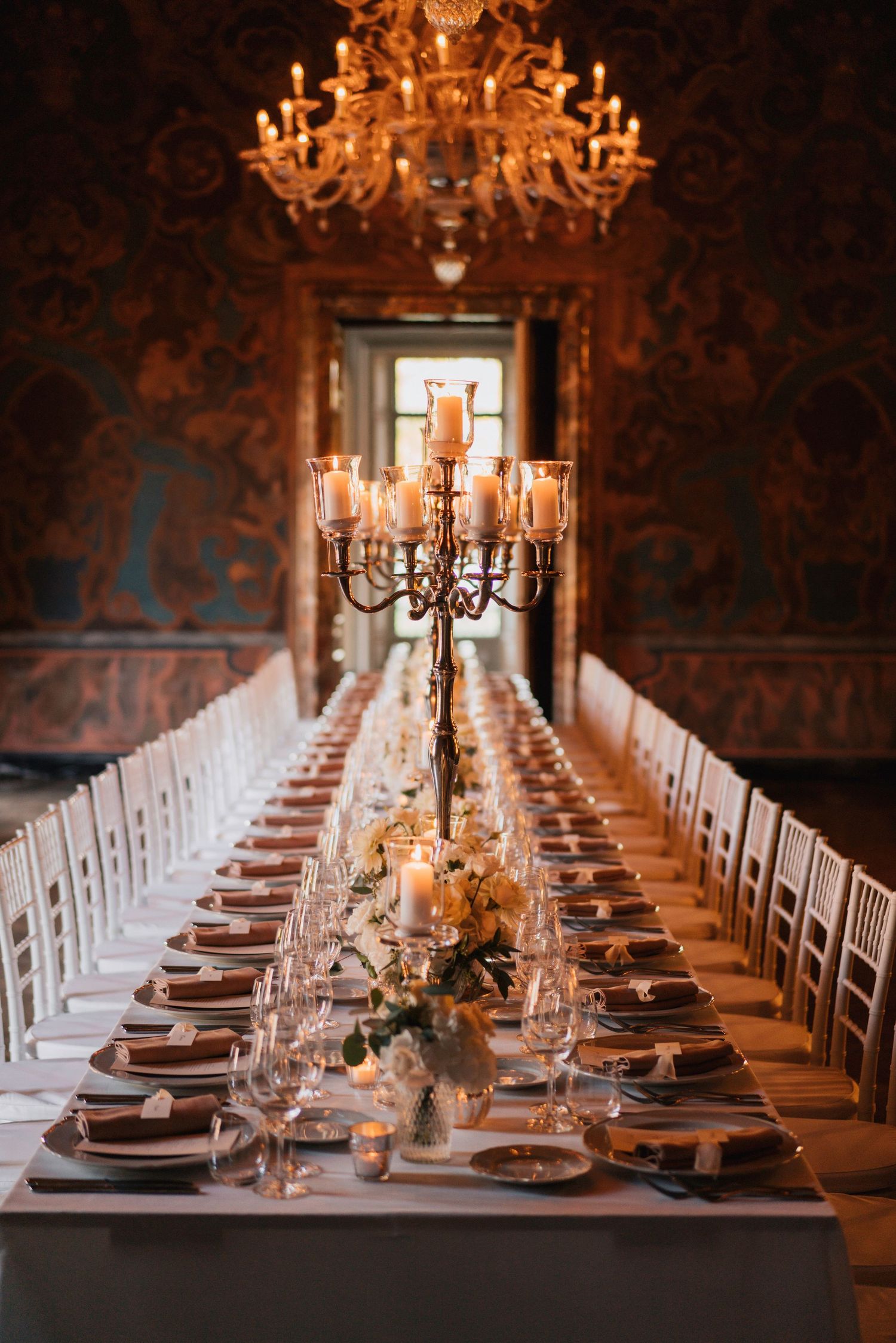 Elegant banquet hall table setup with white chairs, crystal chandeliers, and warm lighting creating a luxurious atmosphere.