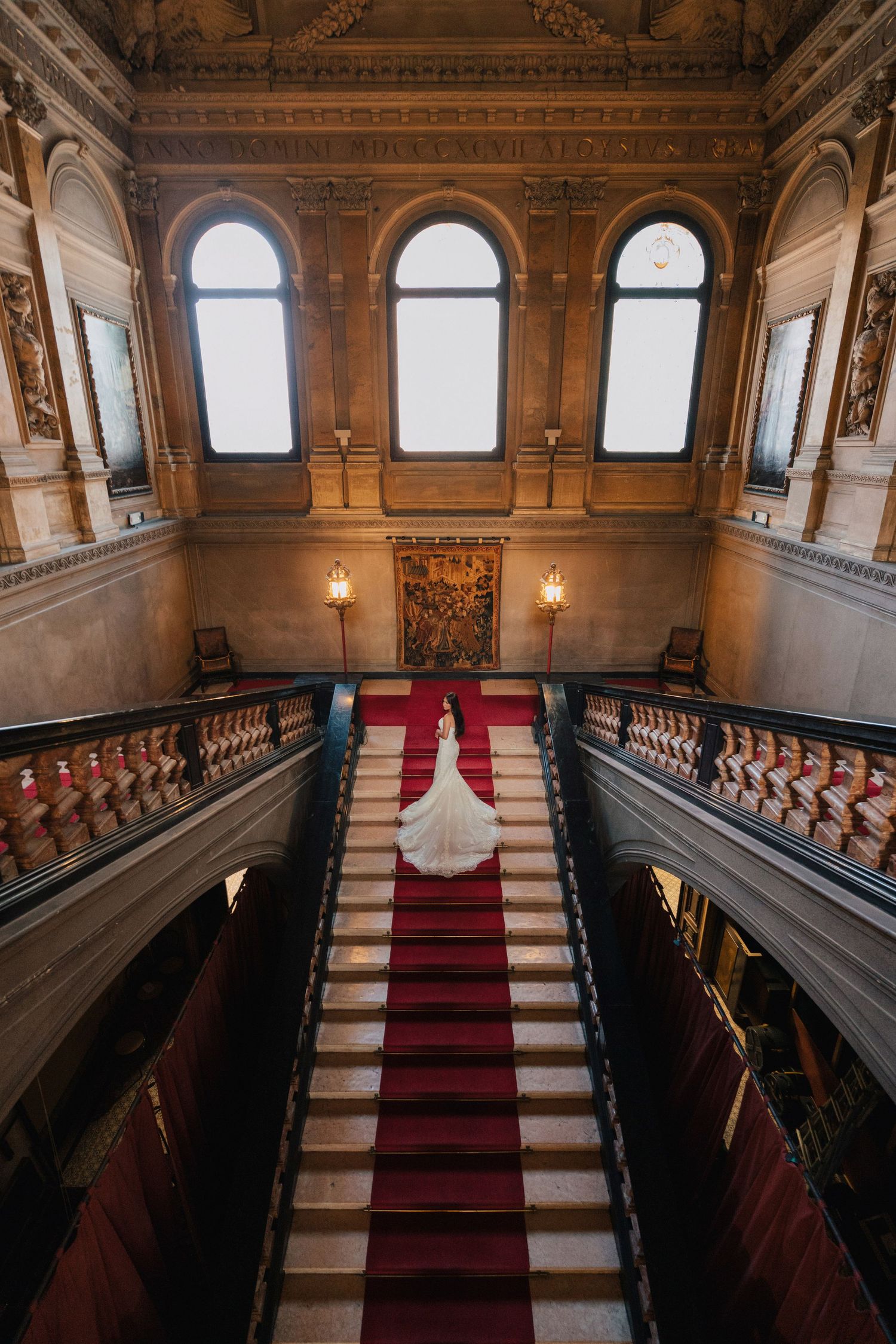A white wedding dress cascades down an ornate grand staircase with red carpet in a luxurious classical building interior.