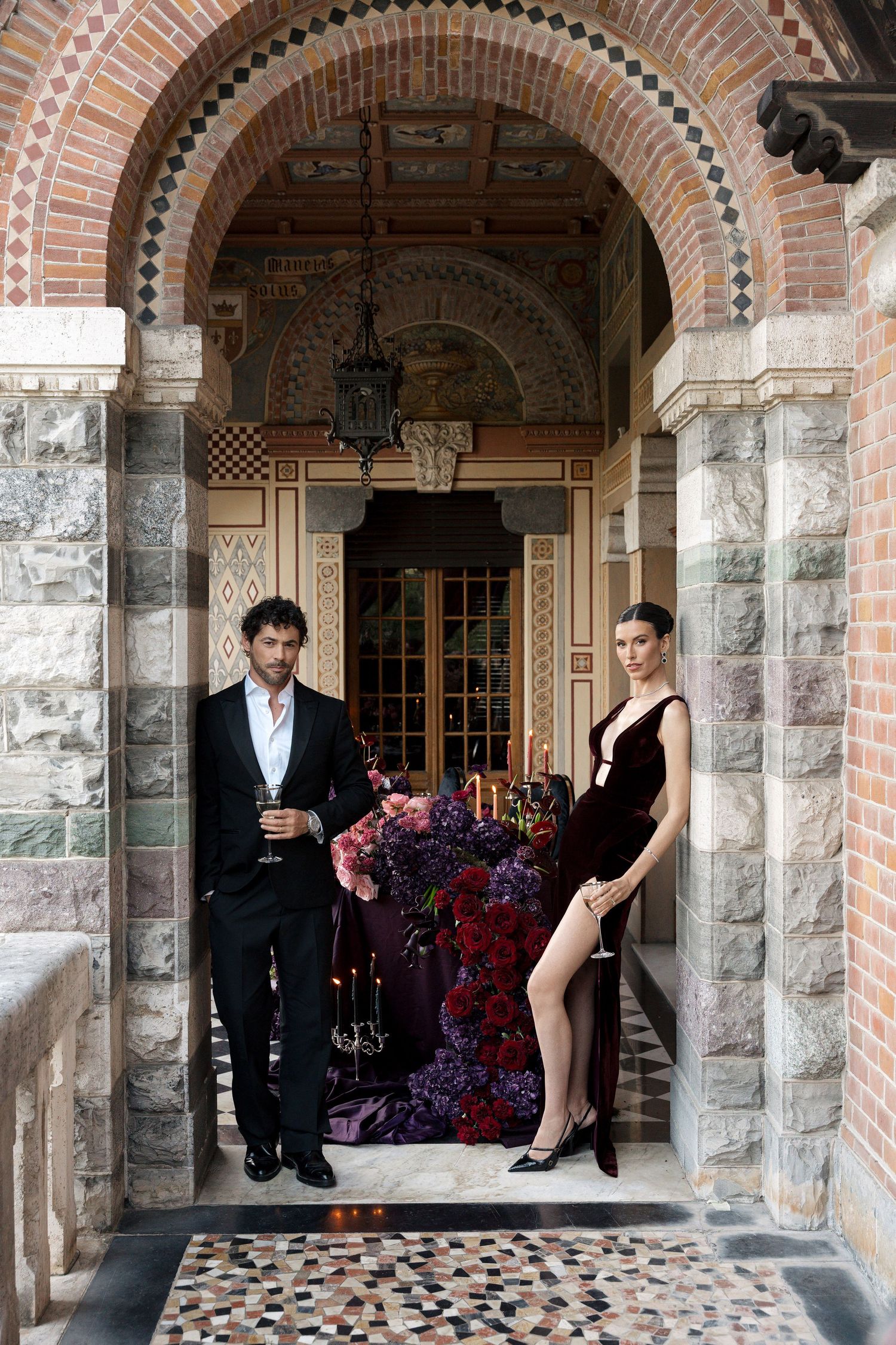 Elegant couple in formal attire standing under ornate brick arch with lush floral arrangement and candles.