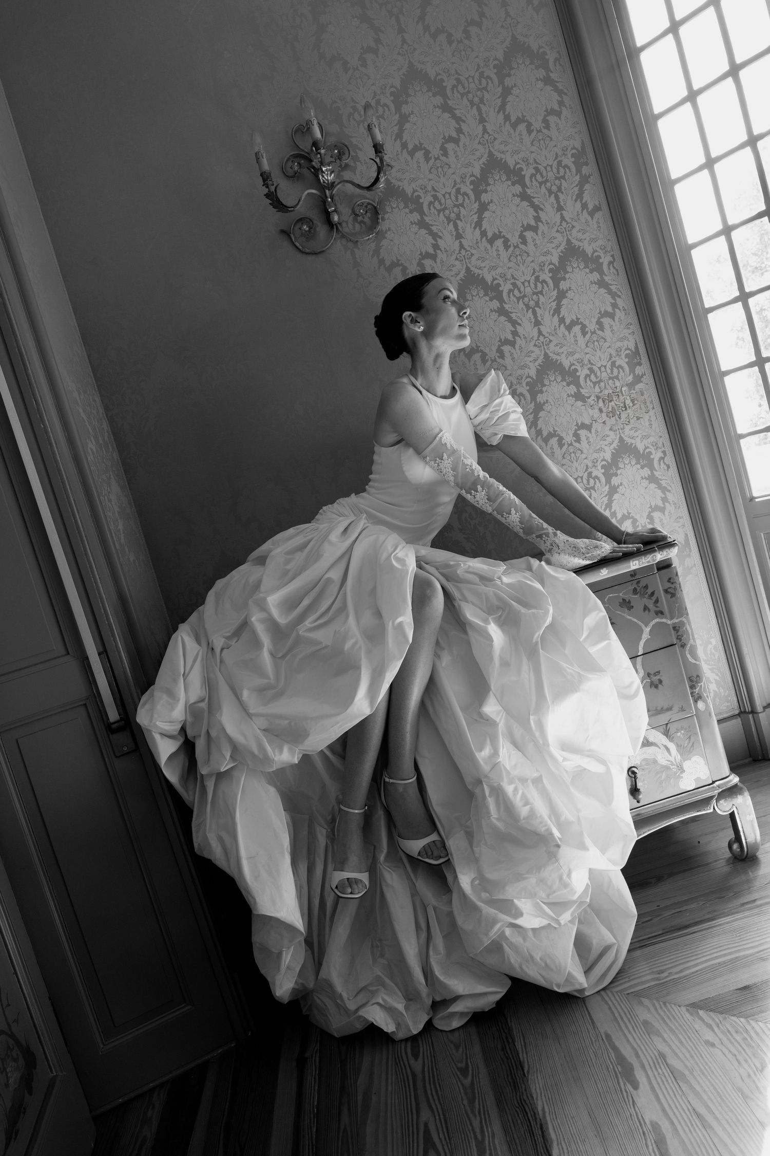 Elegant bride in a voluminous white gown sits gracefully by a window in a vintage room, black and white photo.