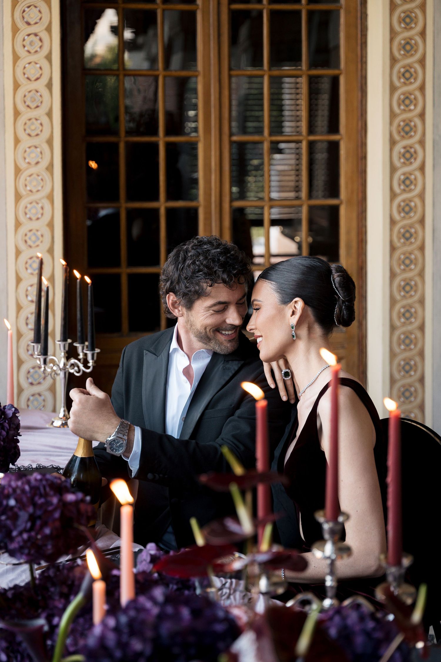 Elegant couple sharing a romantic candlelit dinner, surrounded by purple flowers and candelabras in a luxurious setting.