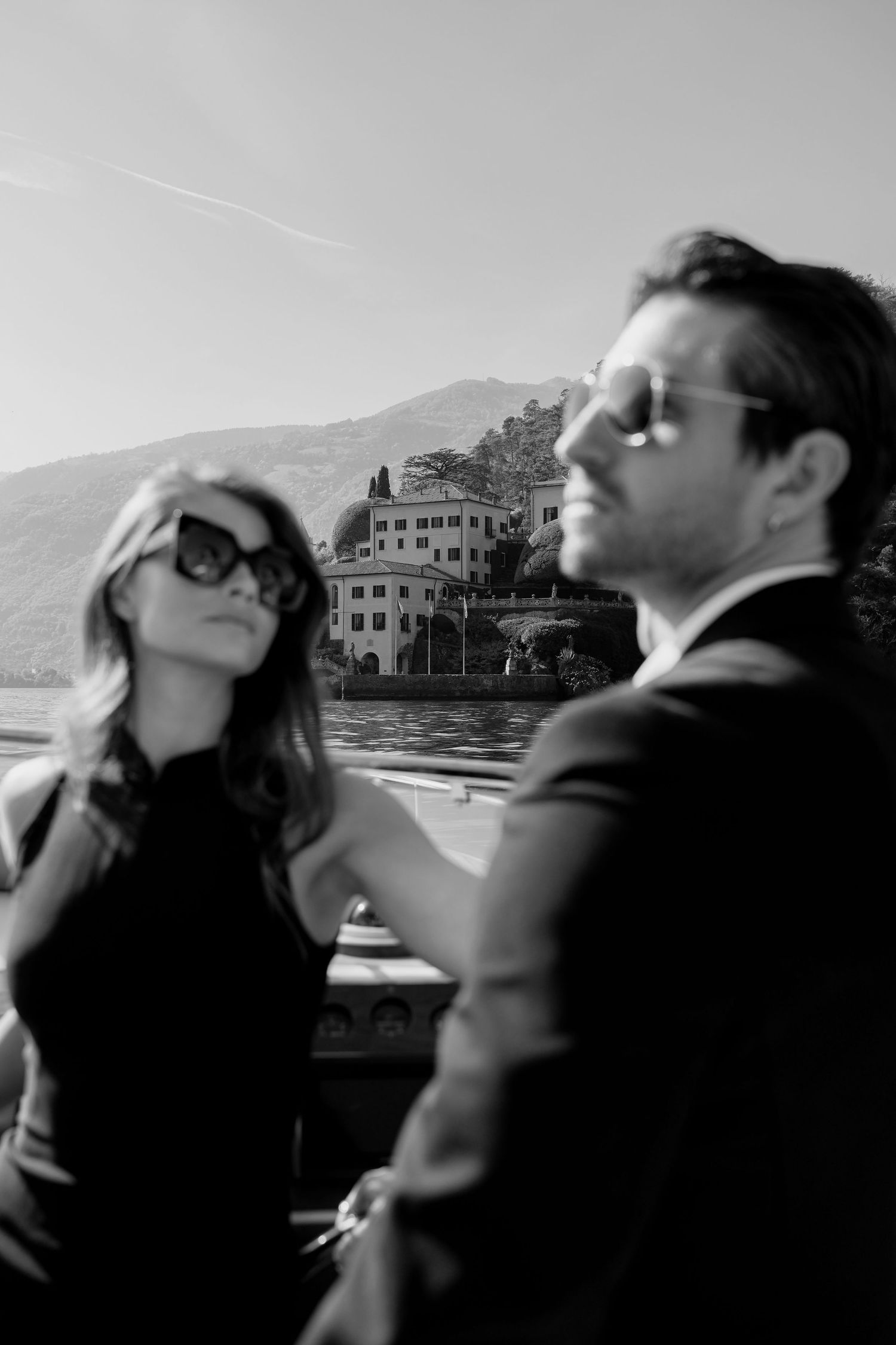 Black and white photo of an elegant couple on a boat with a lakeside villa and mountains in the background.