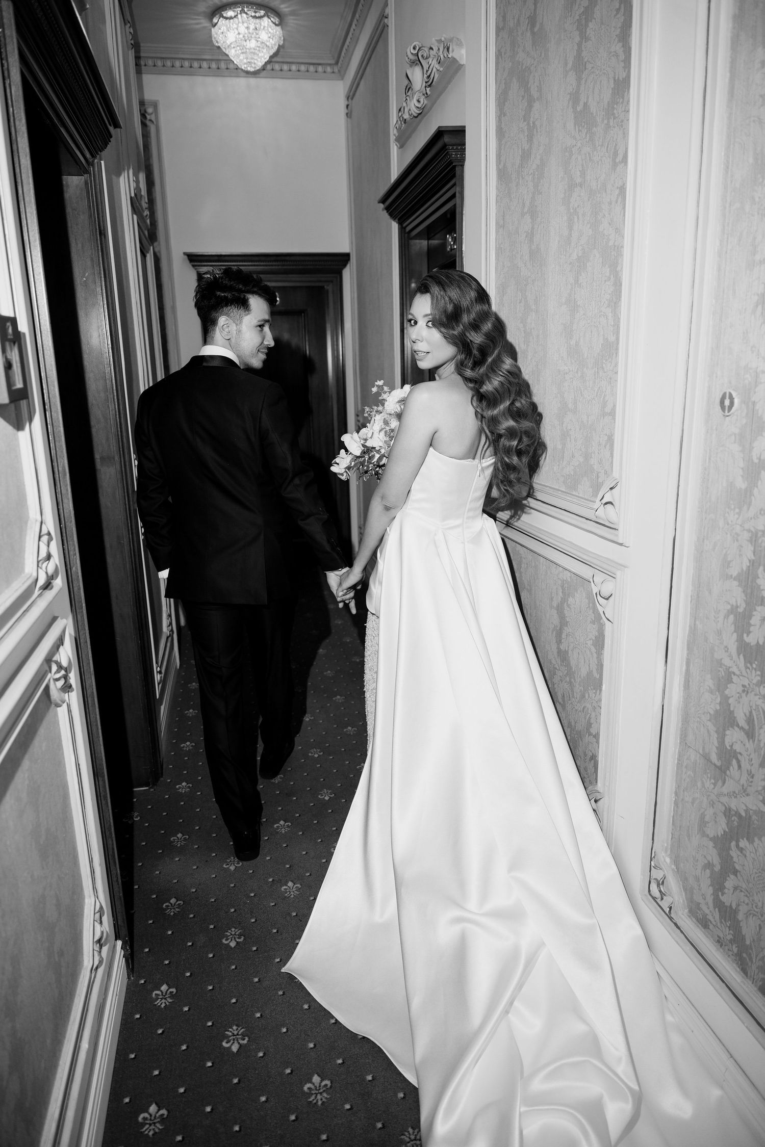 Wedding couple walking down an elegant hallway with marble walls and crystal chandeliers.