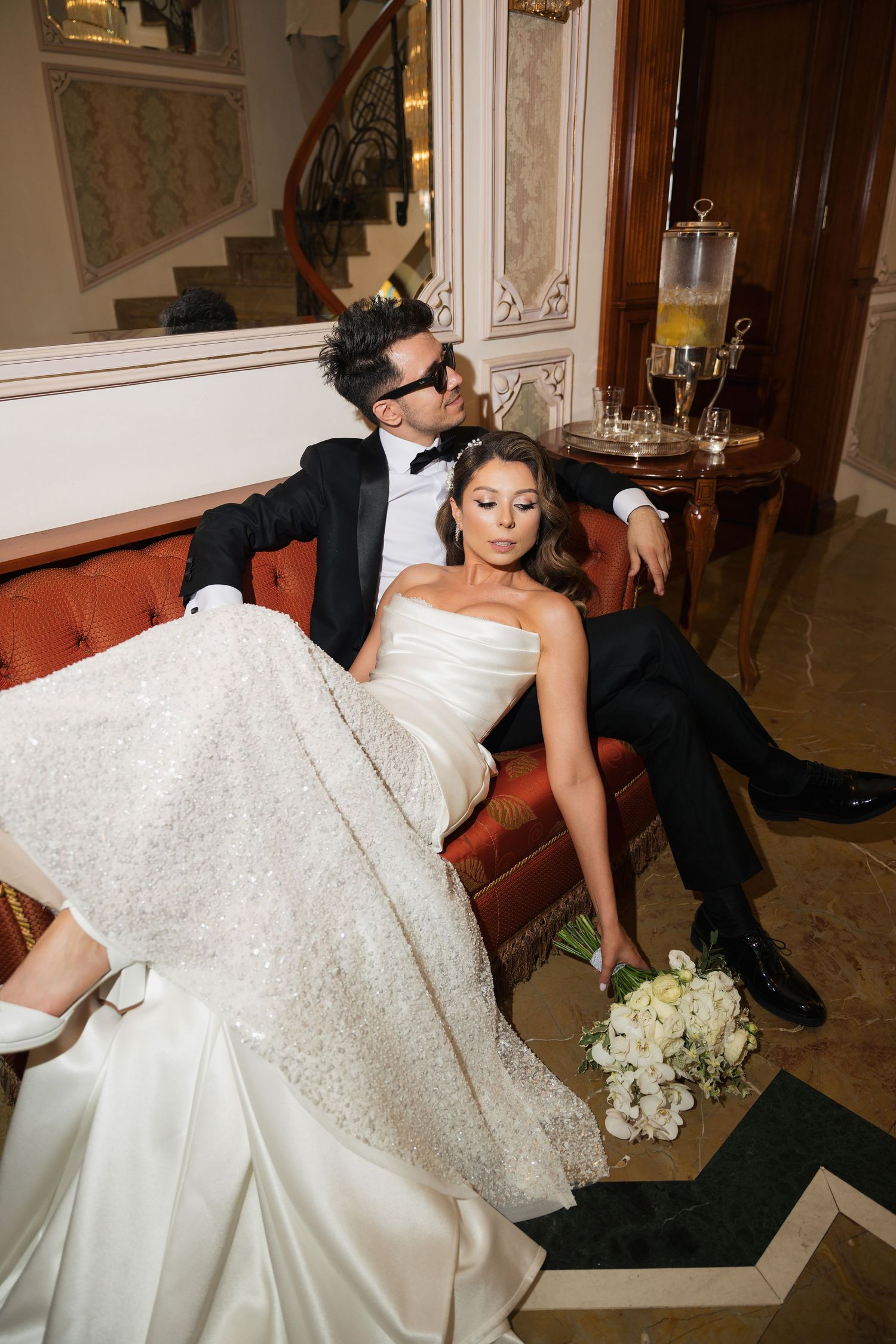 Bride in luxurious white gown reclining on antique furniture during wedding photoshoot.