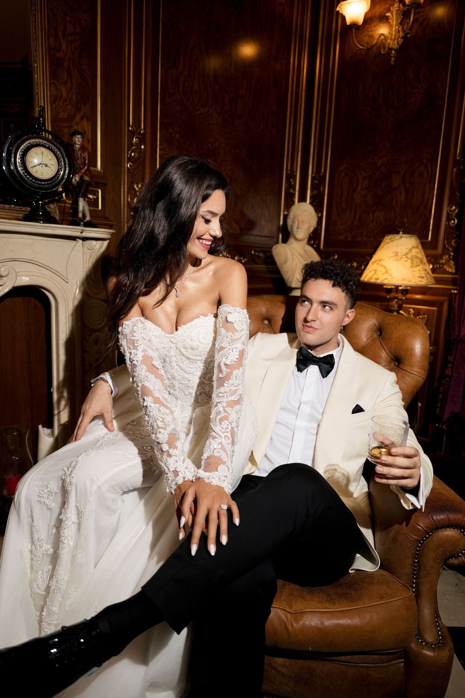 Elegant couple in wedding attire lounging on a vintage leather chair in a luxurious room.