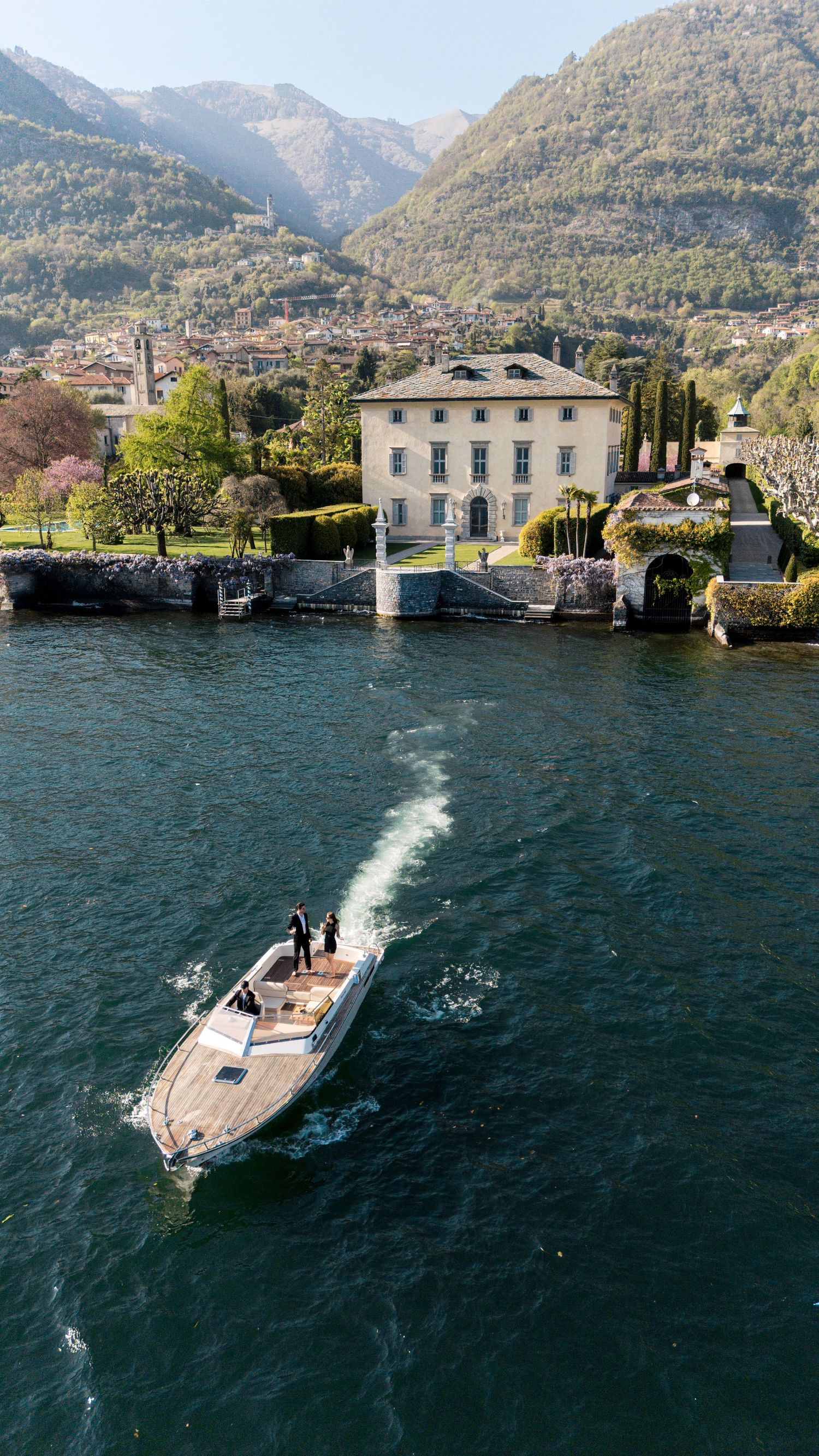 Aerial view of a wooden speedboat on Lake Como with a historic Italian villa and lush mountains in the background.