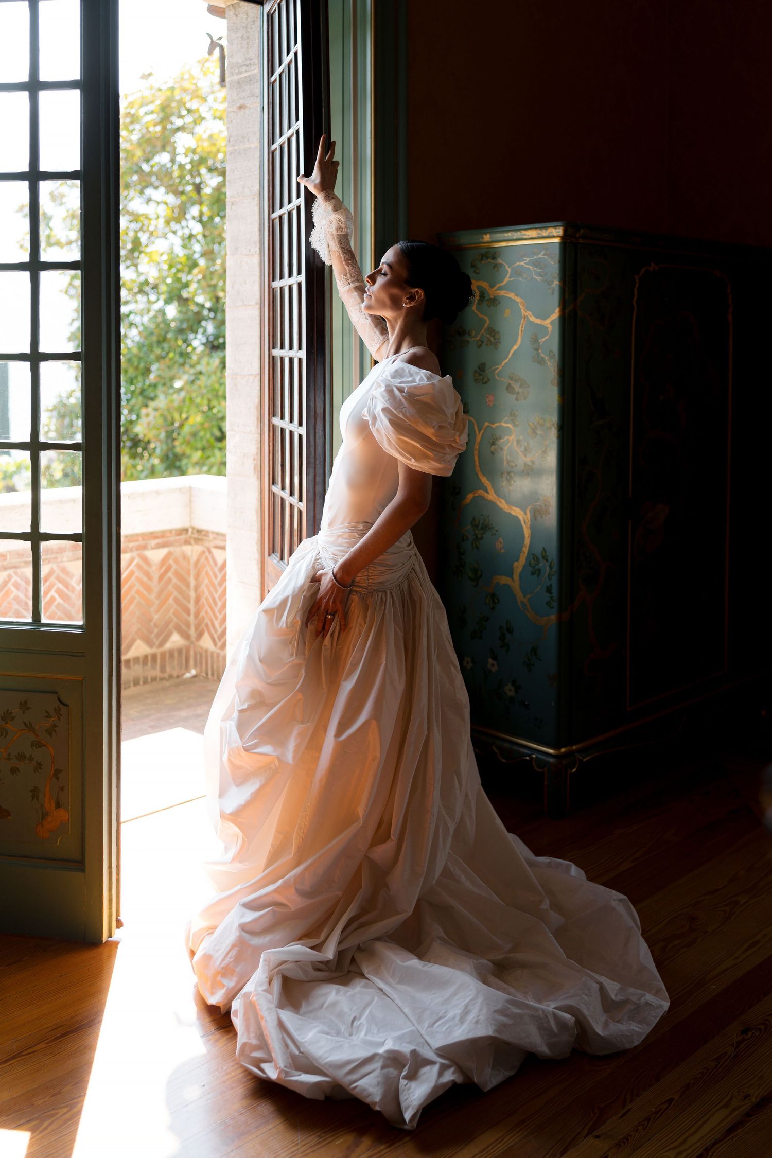 Bride in elegant white gown standing by open French doors, bathed in natural light inside a historic mansion.