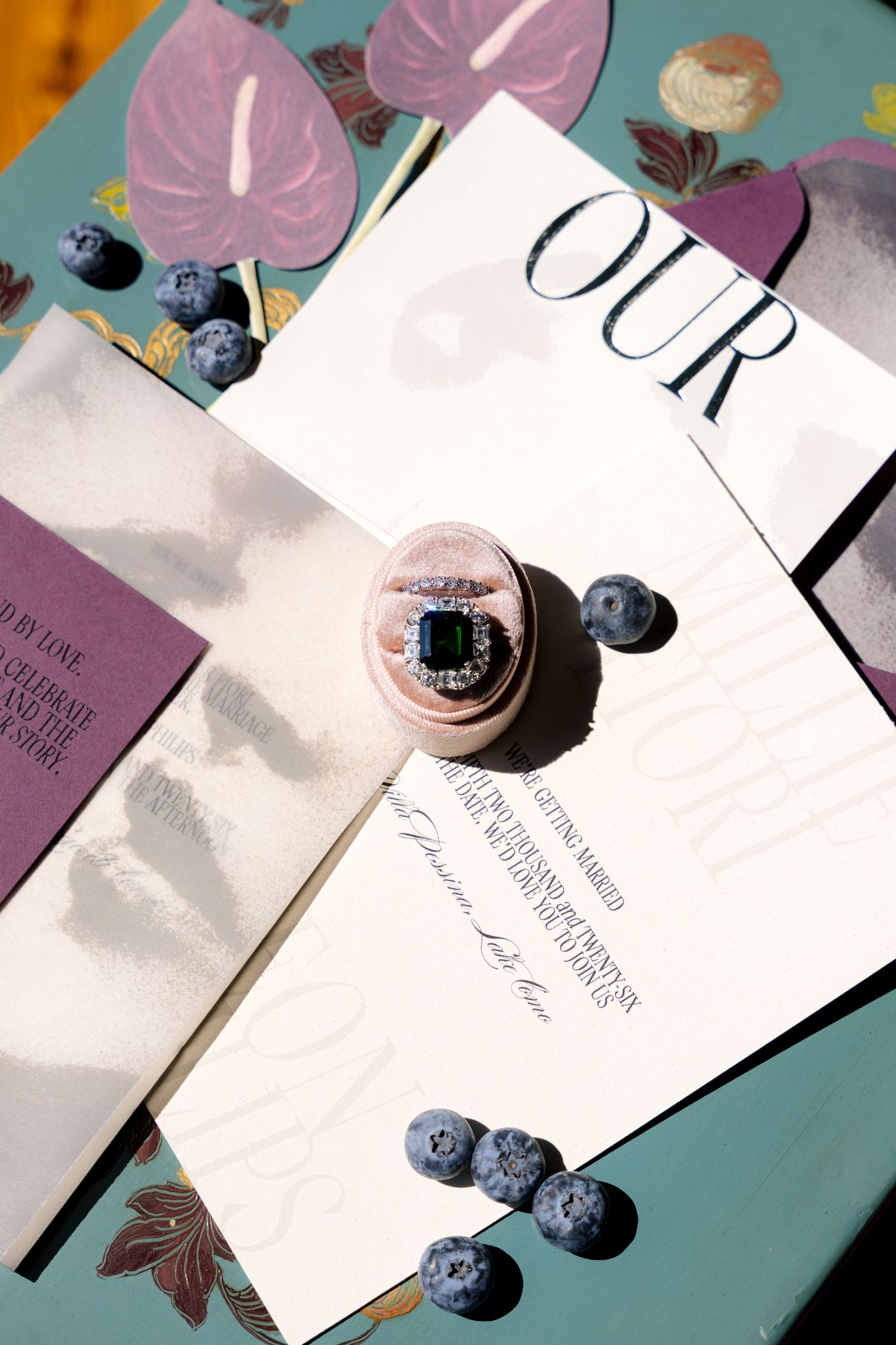 Elegant wedding invitation suite with emerald engagement ring, blueberries, and floral accents on a teal surface.