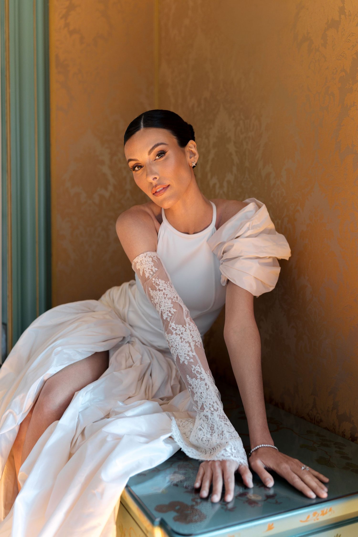 Elegant bride in white gown with lace gloves and ruffled sleeves sits gracefully against a golden wall.