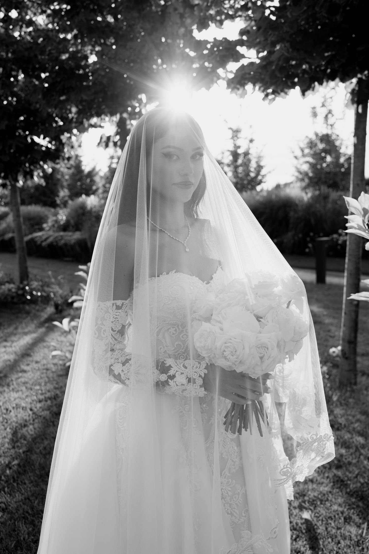 Black and white bridal portrait with sunlight streaming through trees and delicate lace veil.