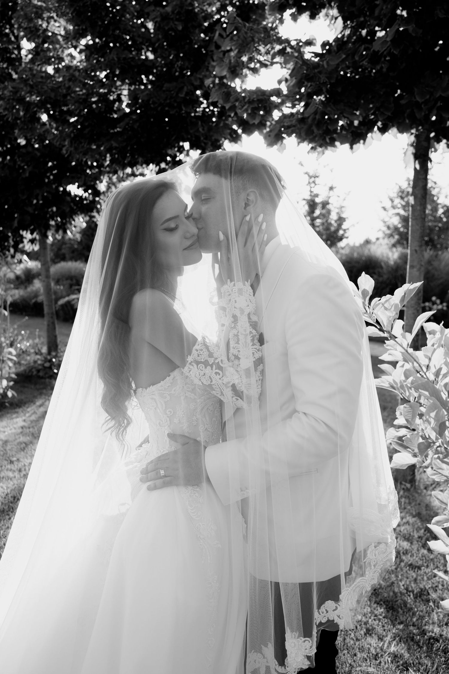 A romantic black and white wedding photo series showing a couple in white embracing outdoors among trees and foliage.