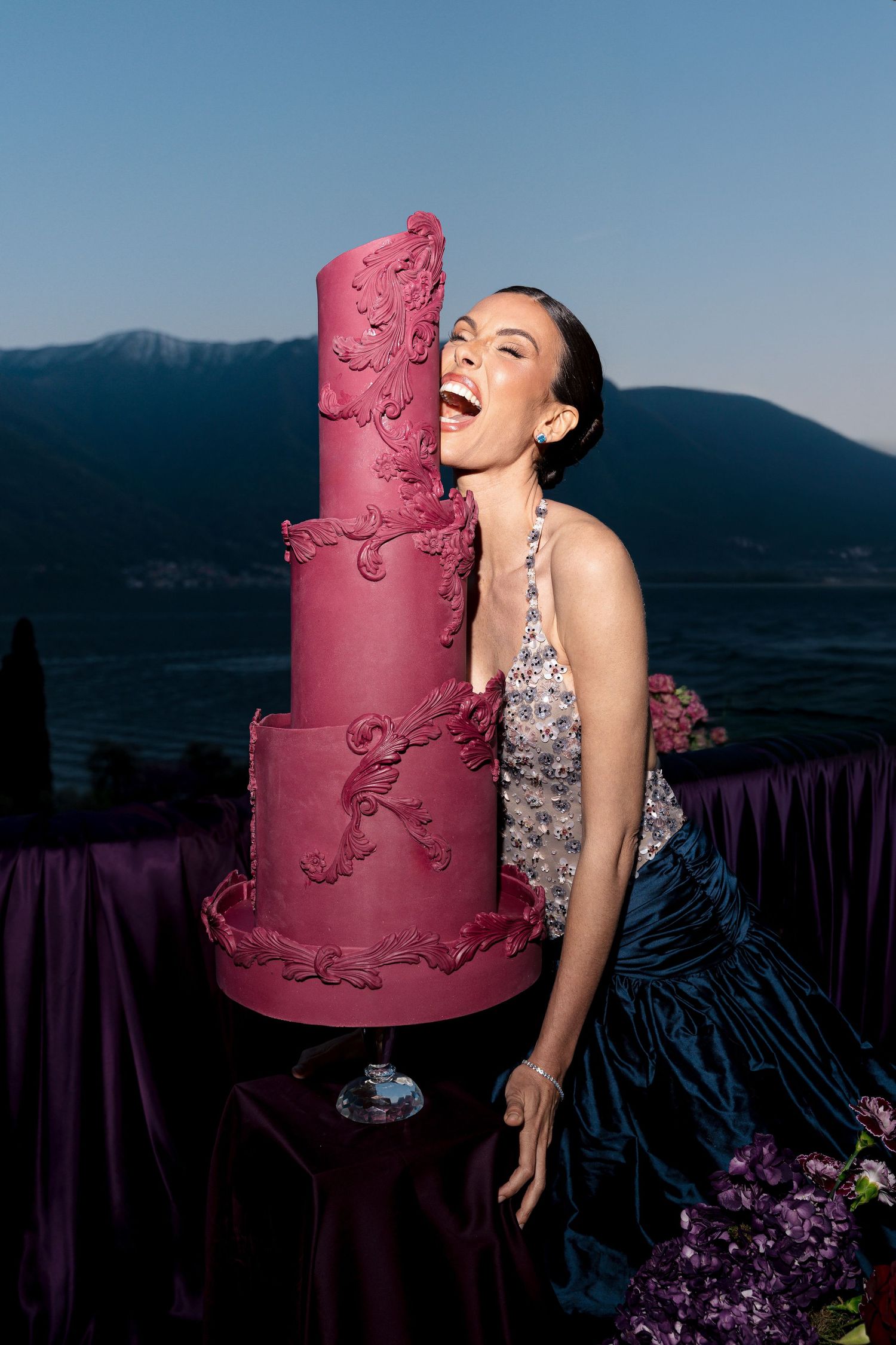 Woman in silver gown laughing beside a tall pink tiered wedding cake with mountain backdrop.