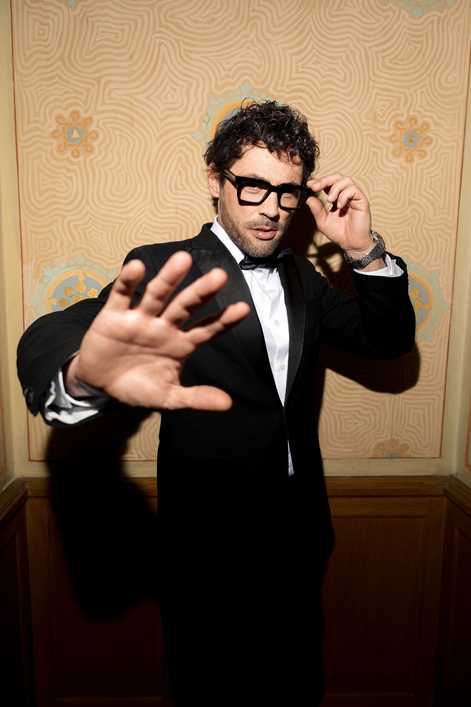 Man in tuxedo and black-rimmed glasses gesturing with hand toward camera in stylish portrait.