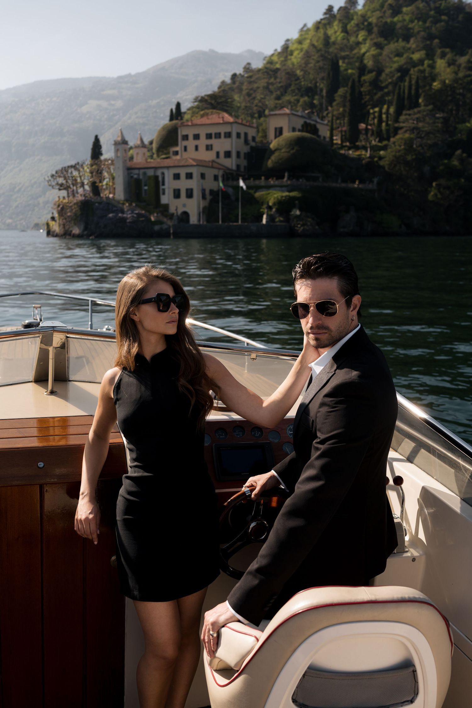 Elegant couple in black outfits and sunglasses on a luxury boat with a scenic Italian lake villa backdrop.