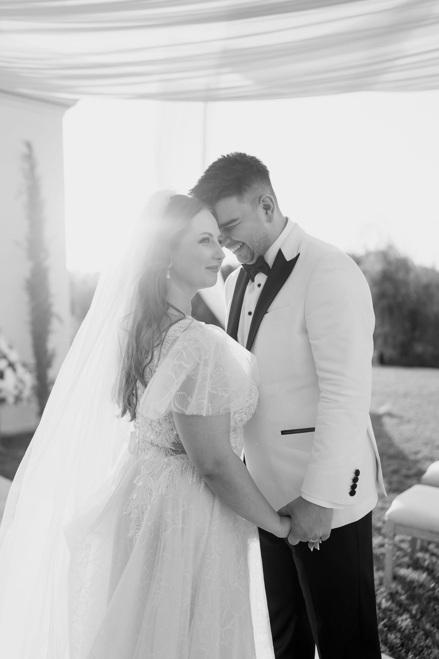 A romantic black and white wedding photo of a couple sharing an intimate moment under draped fabric outdoors.