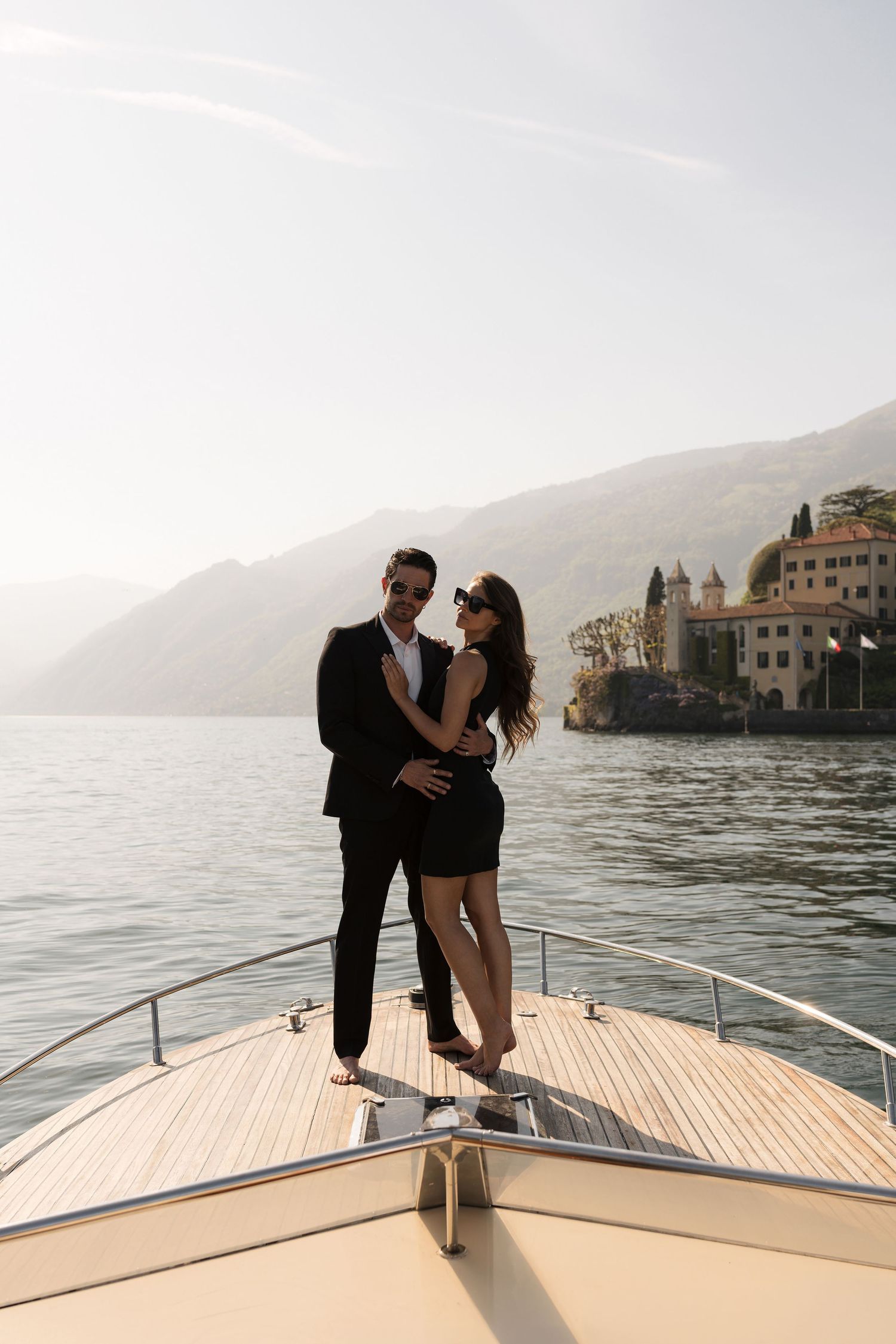 Elegant couple embracing on luxury yacht bow with scenic Lake Como and mountains in background.