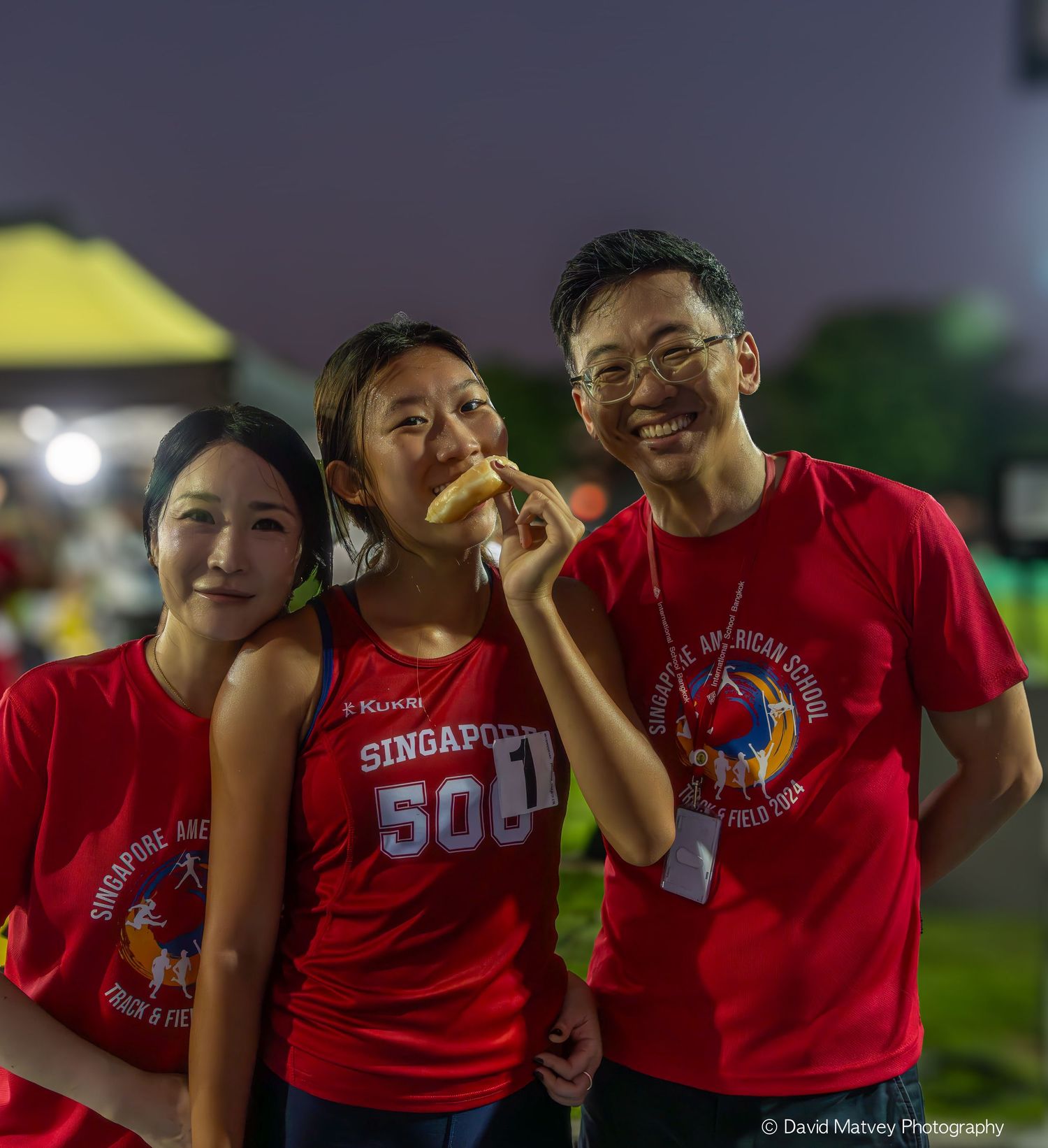 Three people in Singapore American School red shirts smiling at a nighttime event.