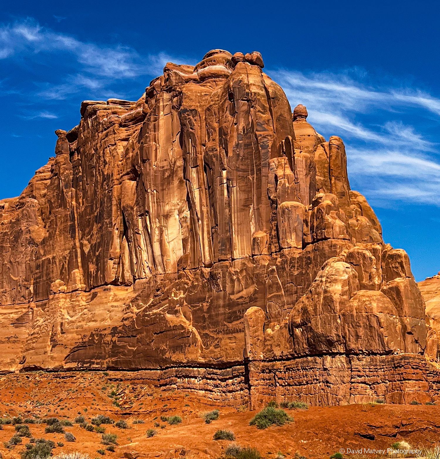 Massive red sandstone butte with dramatic vertical striations under blue sky in Arches National Park.