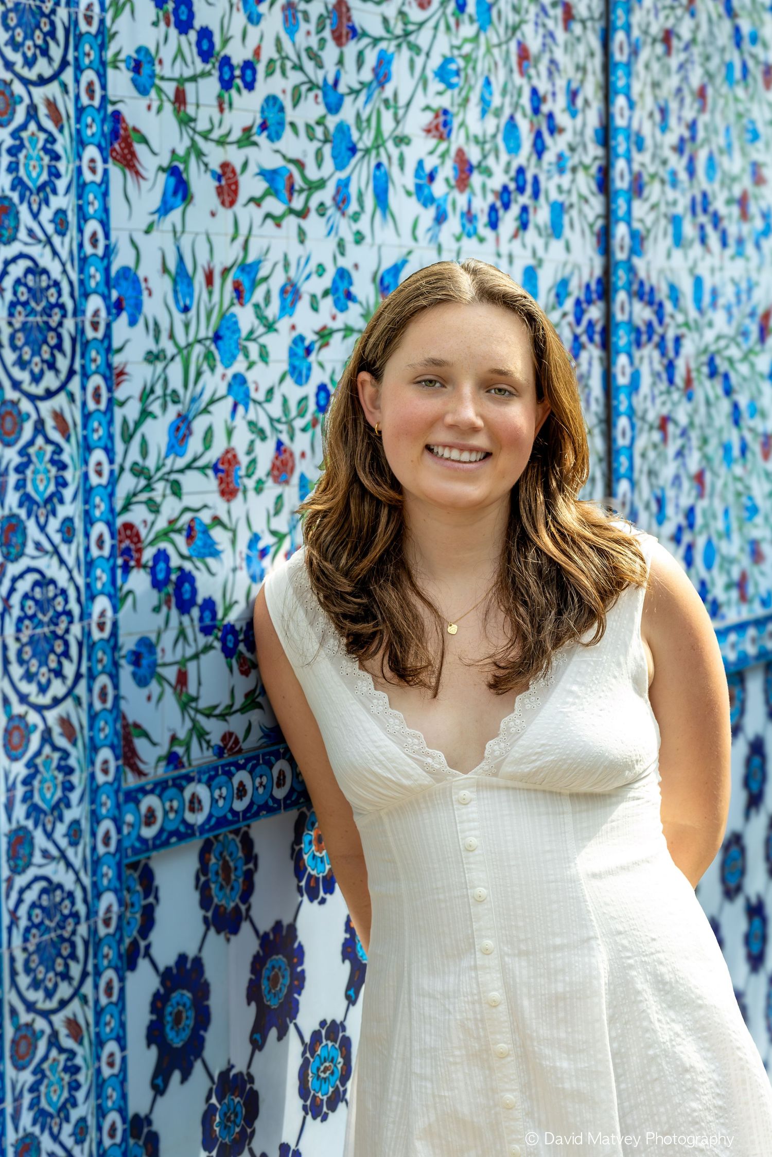 Smiling young woman in white dress leaning against ornate blue floral tile wall.