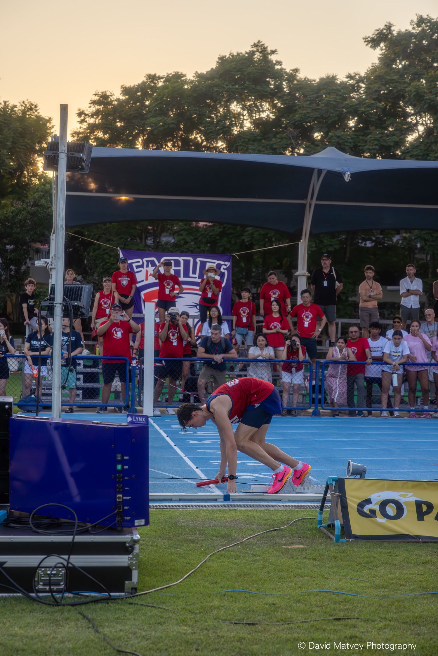 Athlete performs pole vault at outdoor track meet with crowd watching in background at dusk.