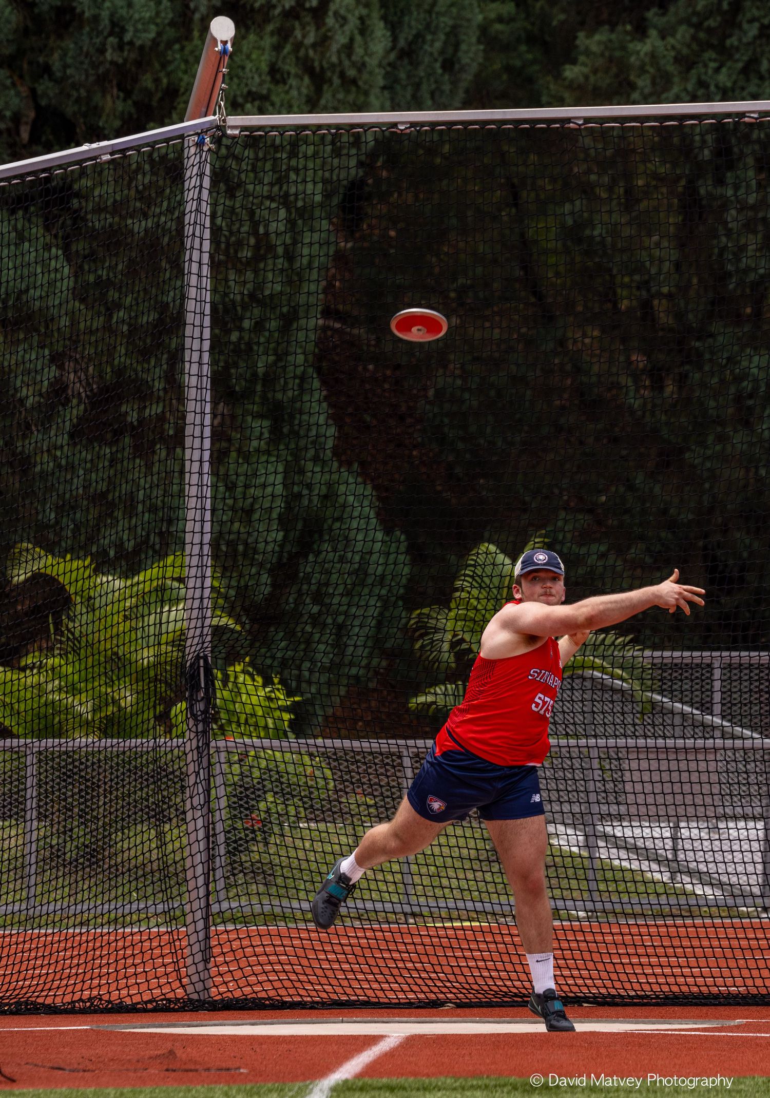 Male athlete in red jersey releases discus throw during outdoor track and field competition.