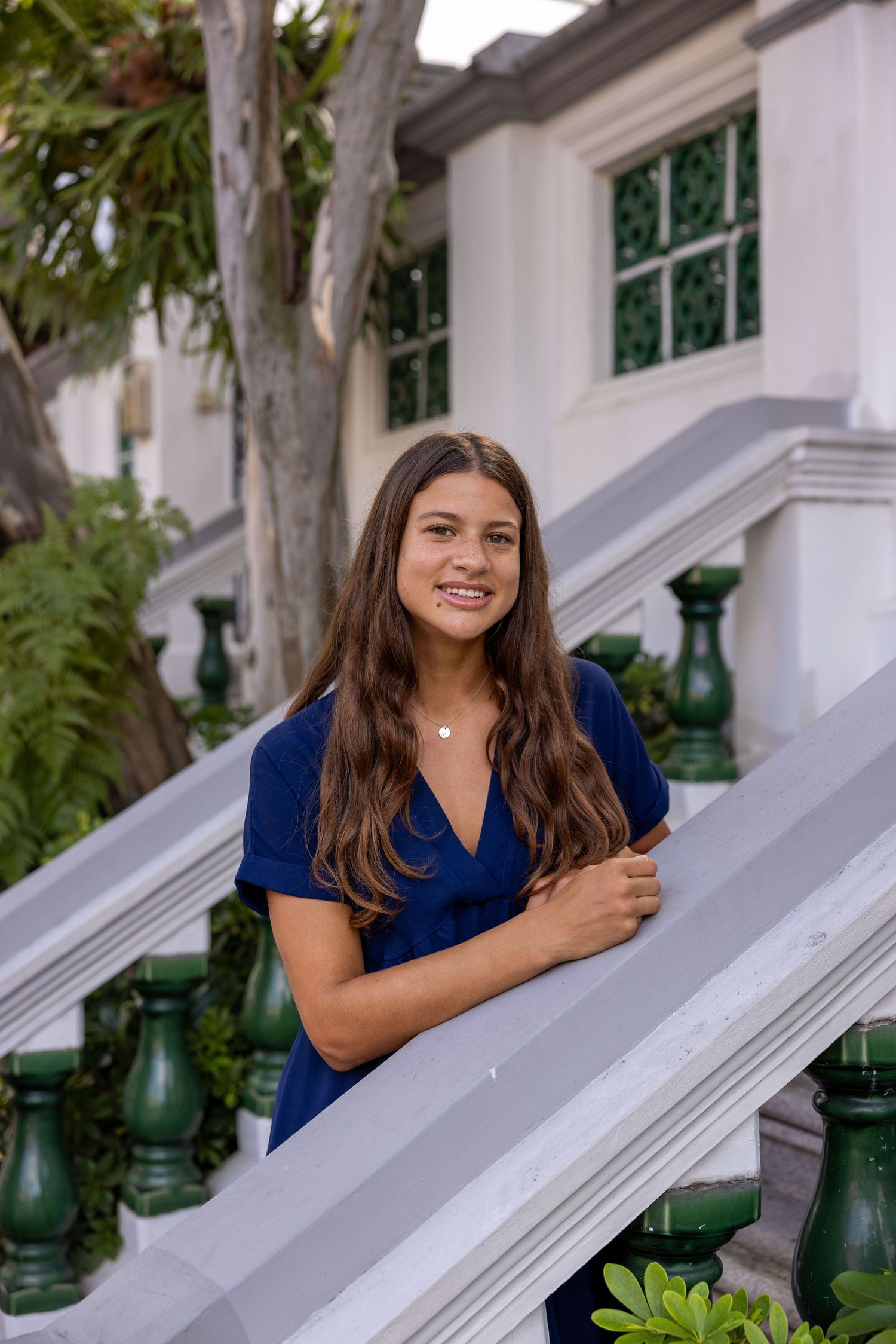 Smiling young woman in blue dress posing on white staircase with green railings and tropical plants.