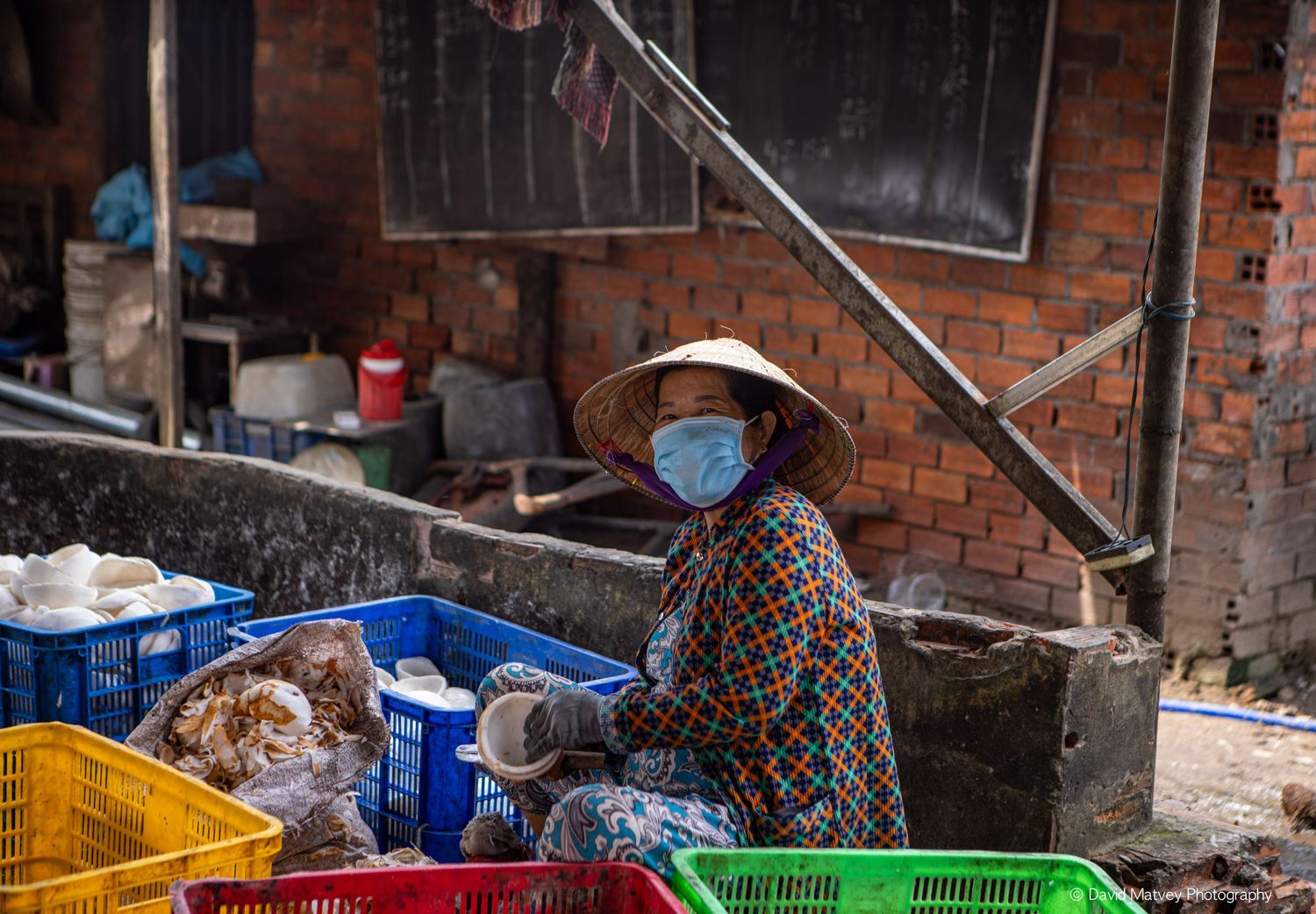 Vietnamese woman wearing conical hat and face mask selling goods at an outdoor market.