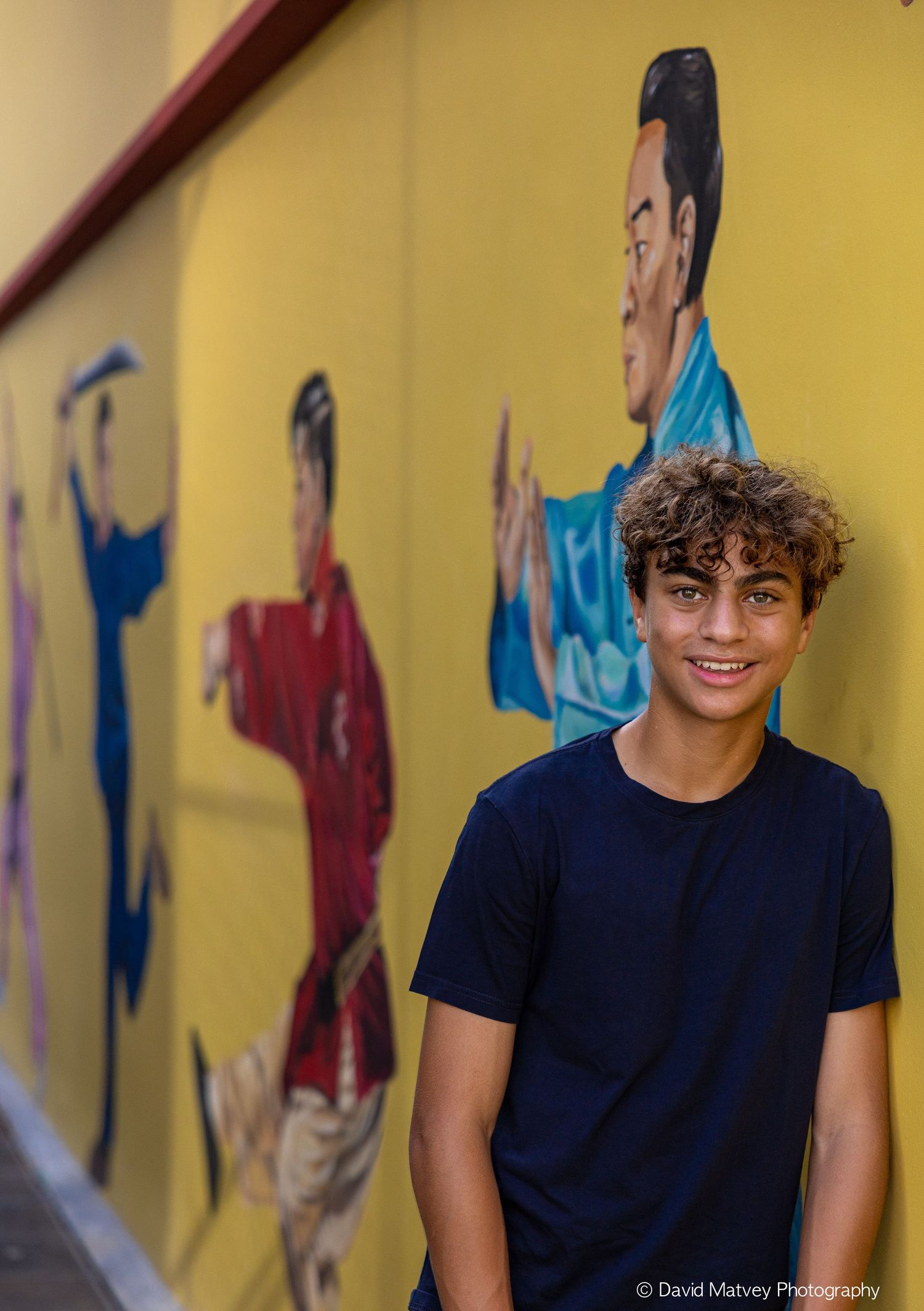 Smiling teenage boy in navy t-shirt standing before a vibrant martial arts street mural.