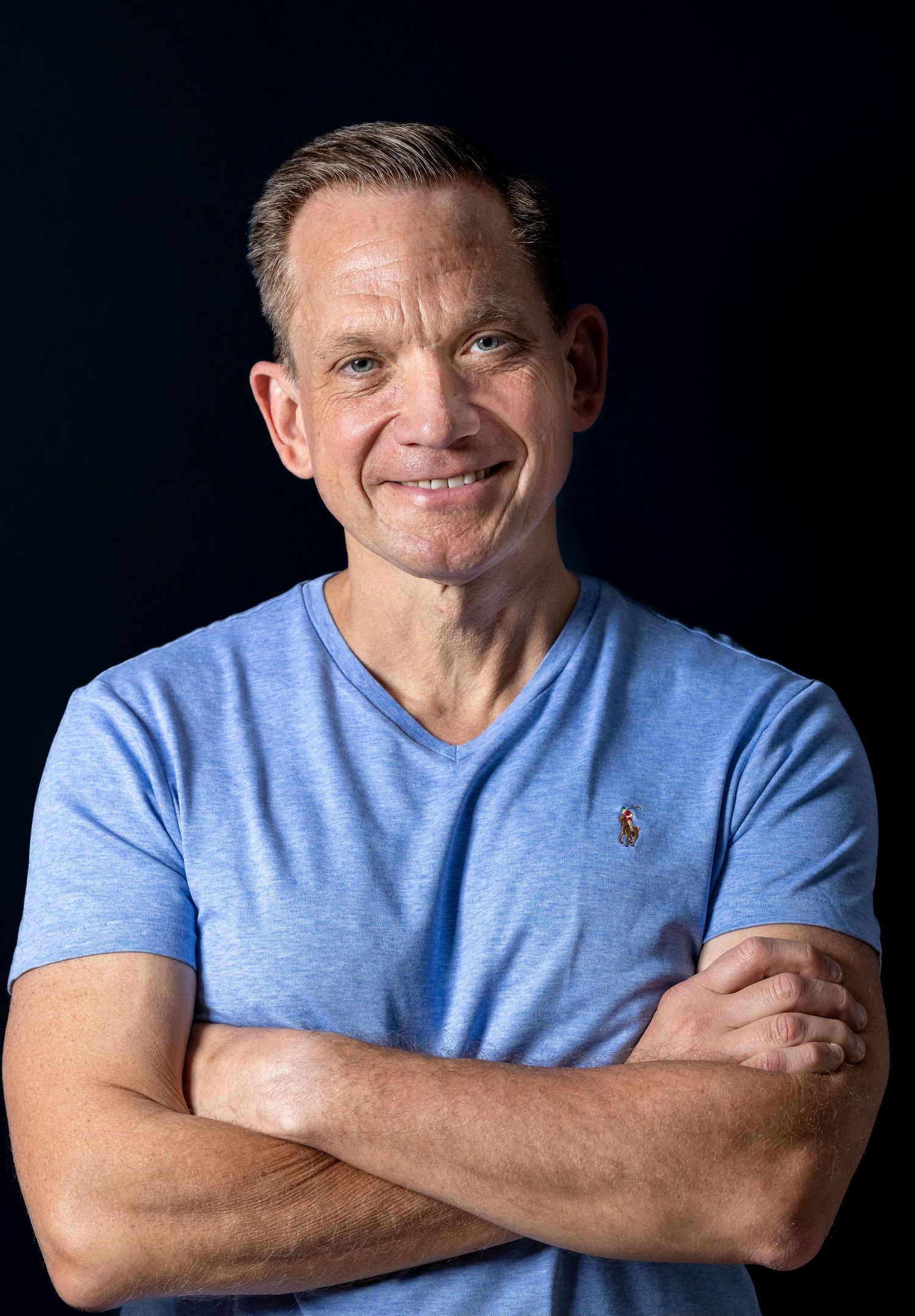 Smiling fit man in blue v-neck t-shirt with arms crossed posing against dark studio background.