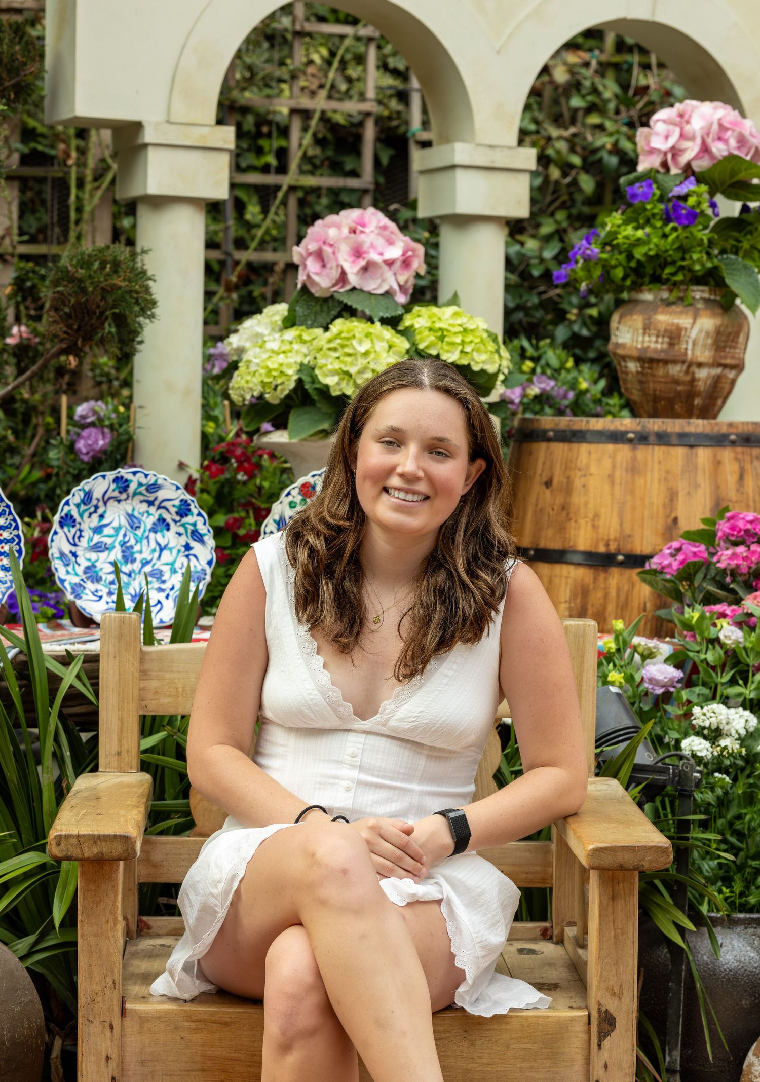 Smiling young woman in white dress seated on wooden bench in colorful flower garden.