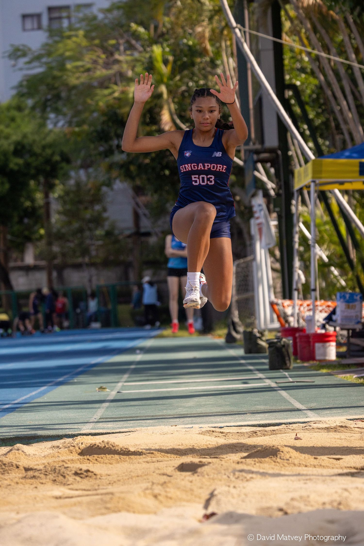 Singapore athlete #503 mid-air during long jump competition on sandy track field.