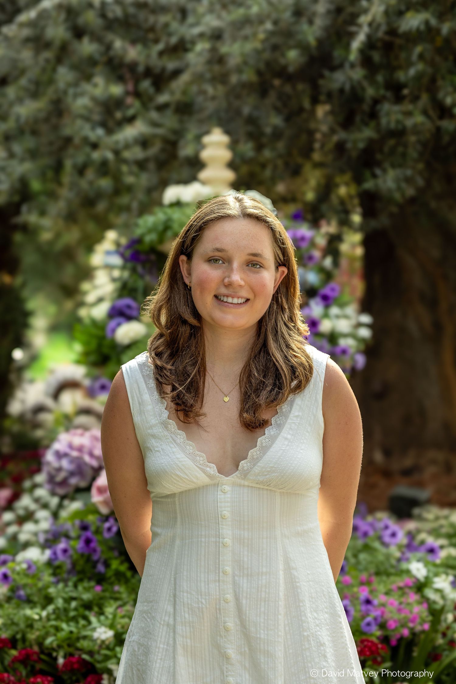 Young woman in white sundress smiling in a colorful garden with purple and white flowers.