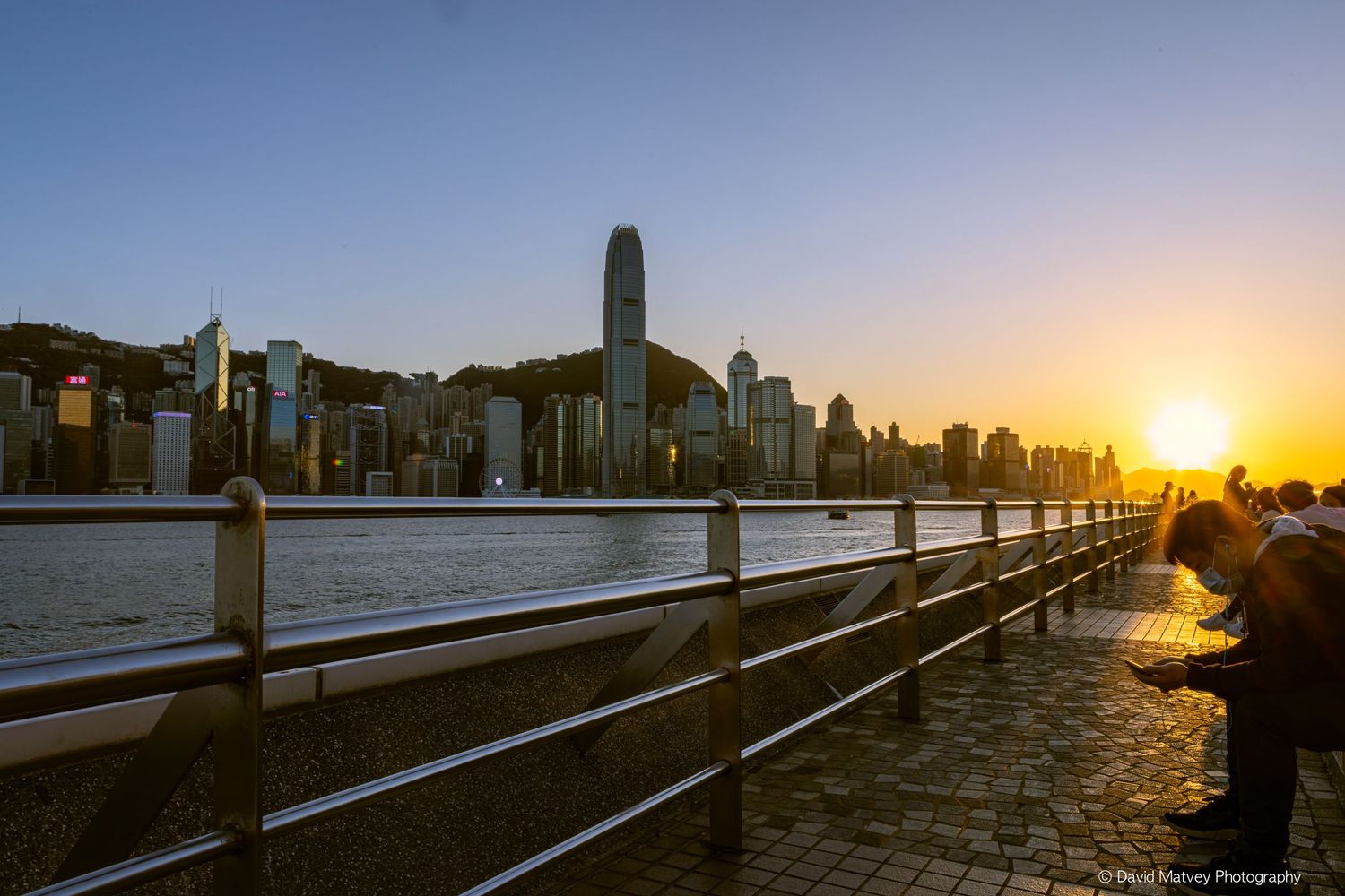Golden sunset over Hong Kong skyline viewed from waterfront promenade.