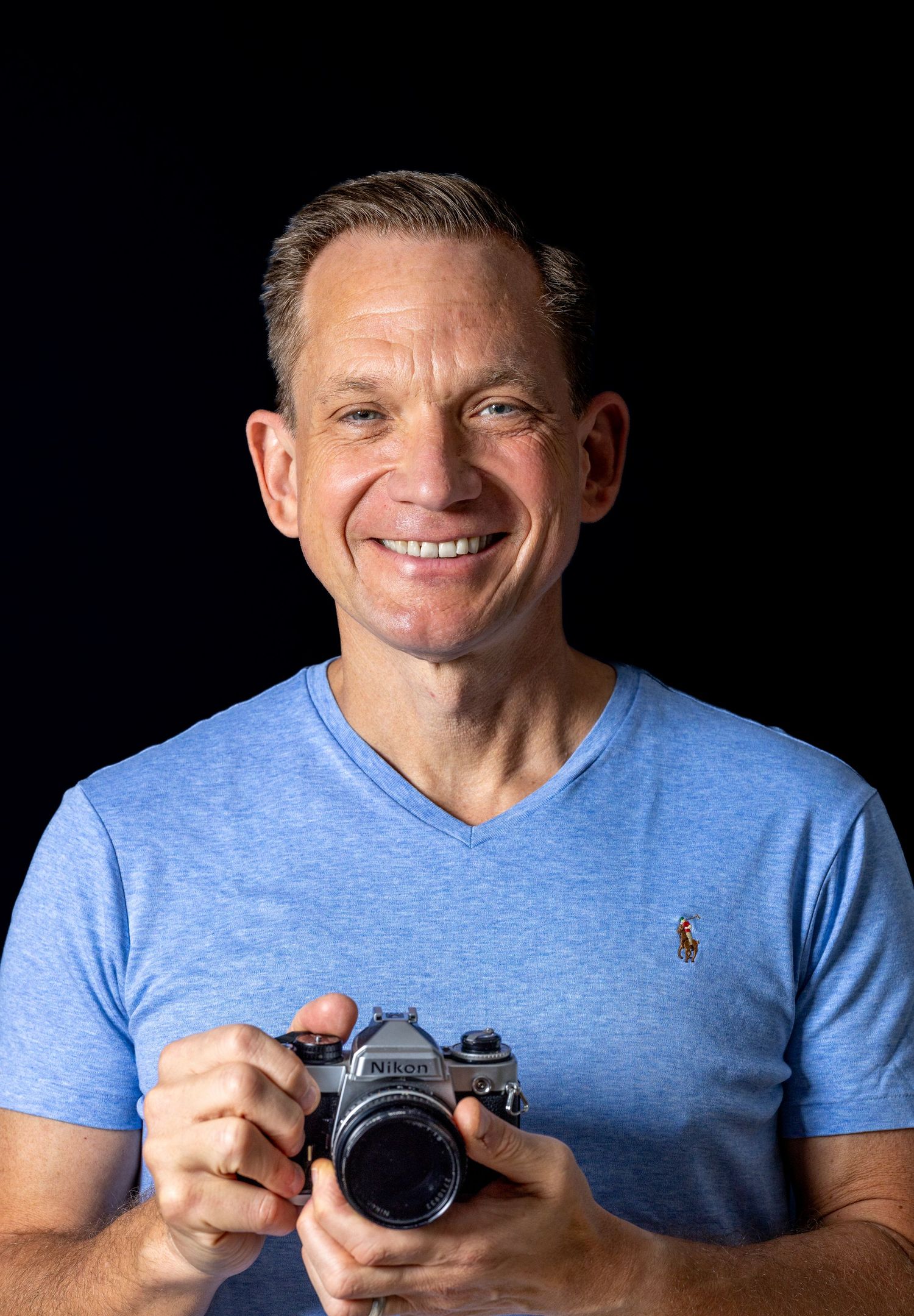 Smiling man in blue t-shirt holding vintage Nikon camera against black studio background.