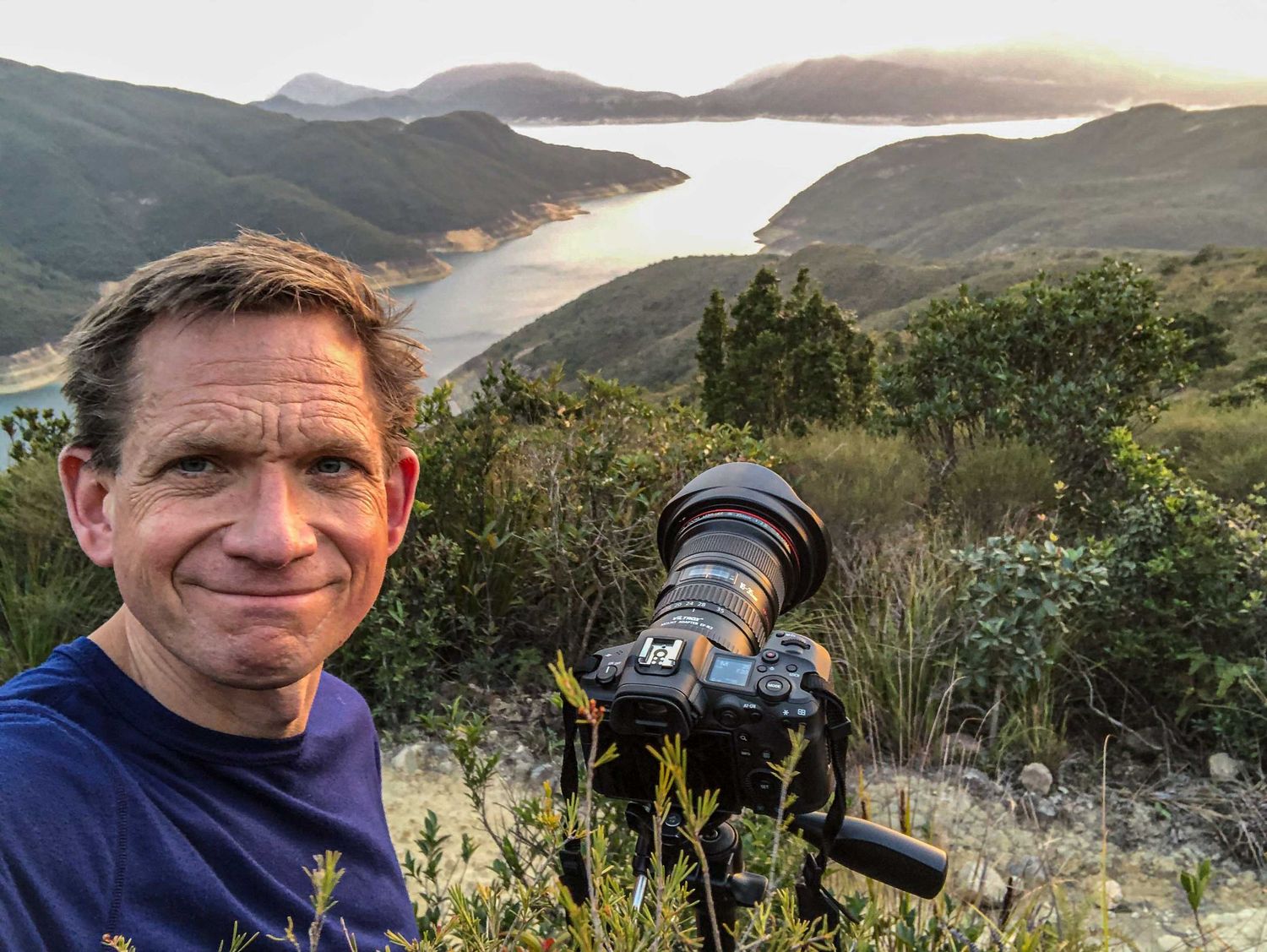 Photographer smiling at camera with telephoto lens on tripod overlooking scenic coastal inlet at golden hour.