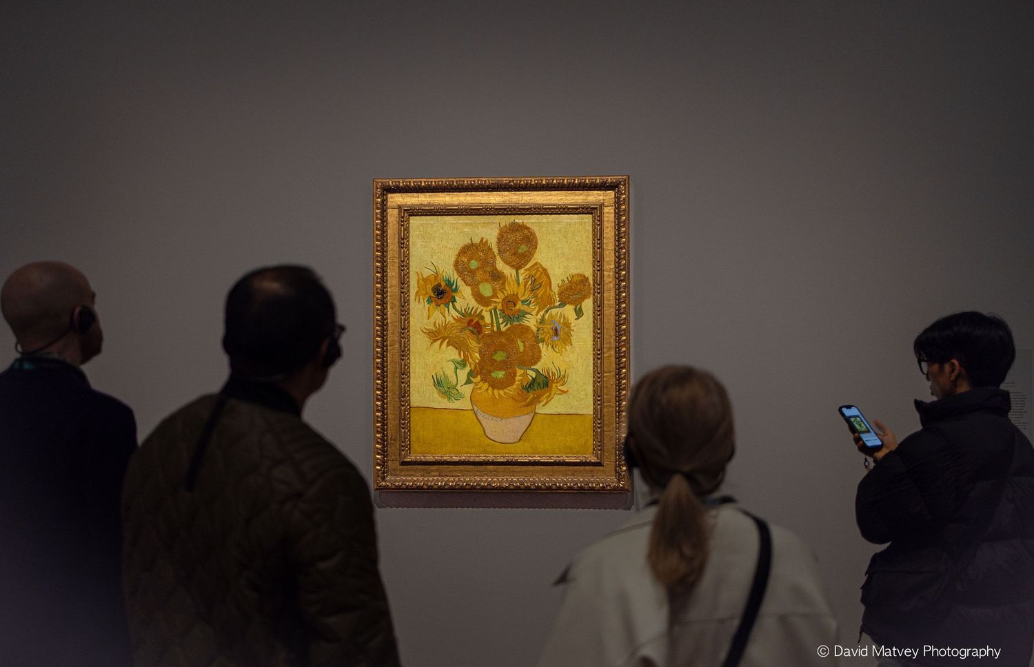 Museum visitors admiring Van Gogh's Sunflowers painting displayed on a gray gallery wall.