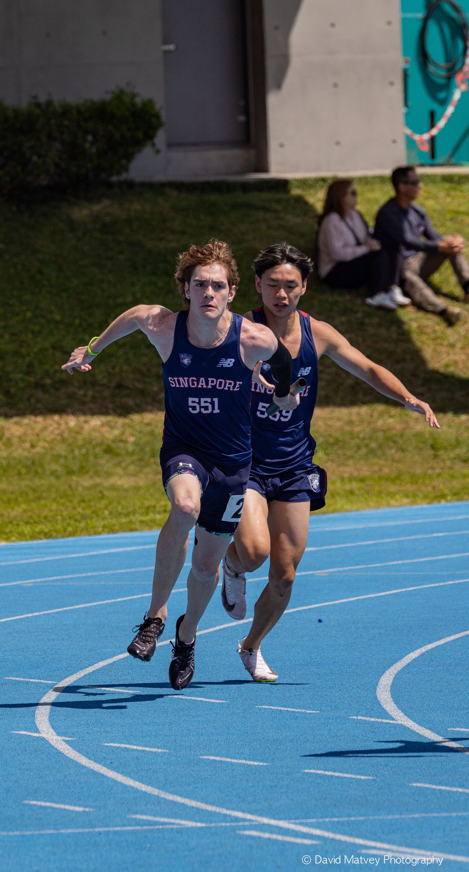 Two Singapore track athletes #551 and #539 sprint during relay race on blue track.
