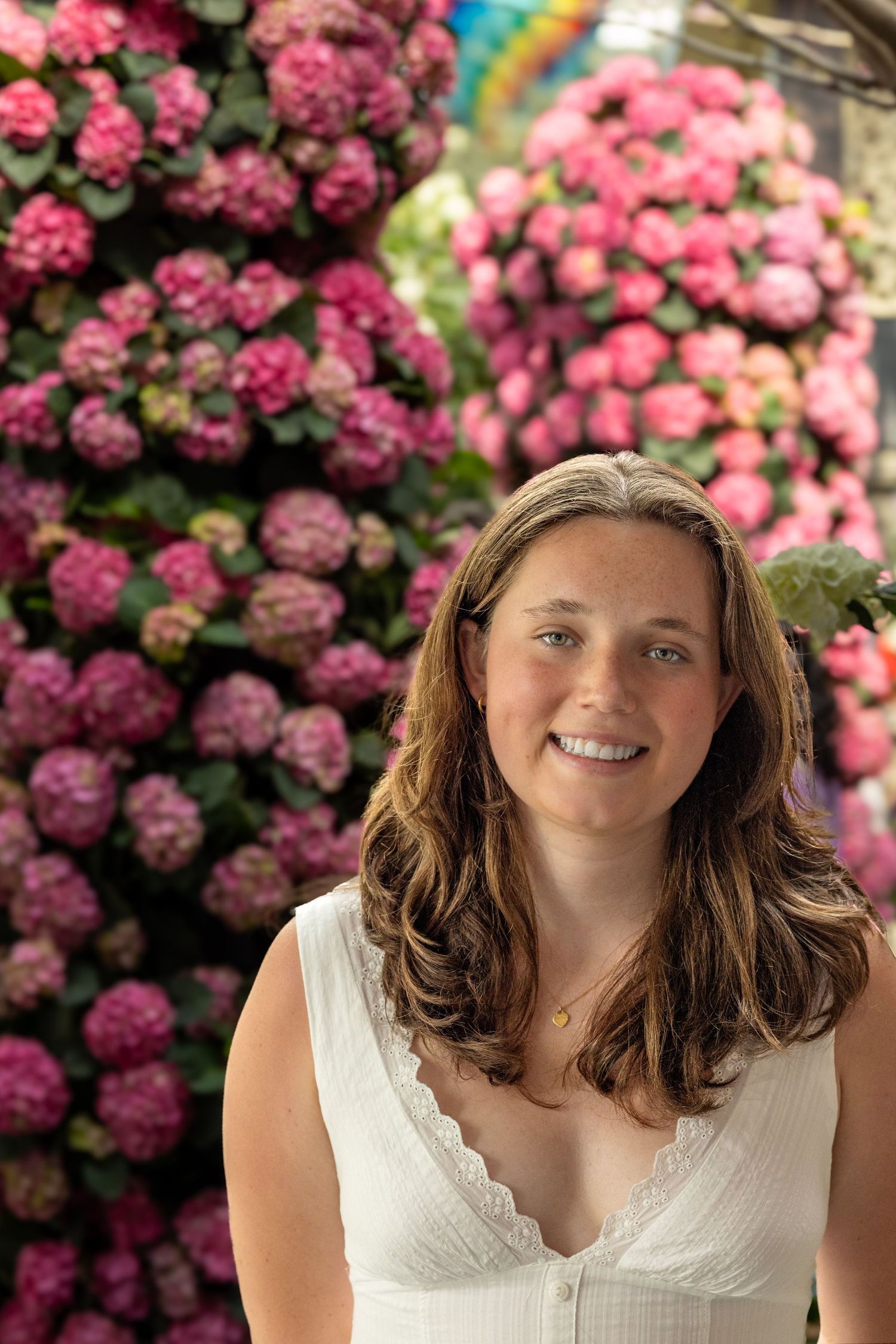 Smiling woman with brown hair in white dress surrounded by pink hydrangea flowers.