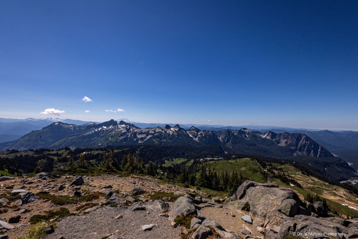 Panoramic summit view of Cascade mountain range with rocky foreground and vast forested valleys below blue sky.