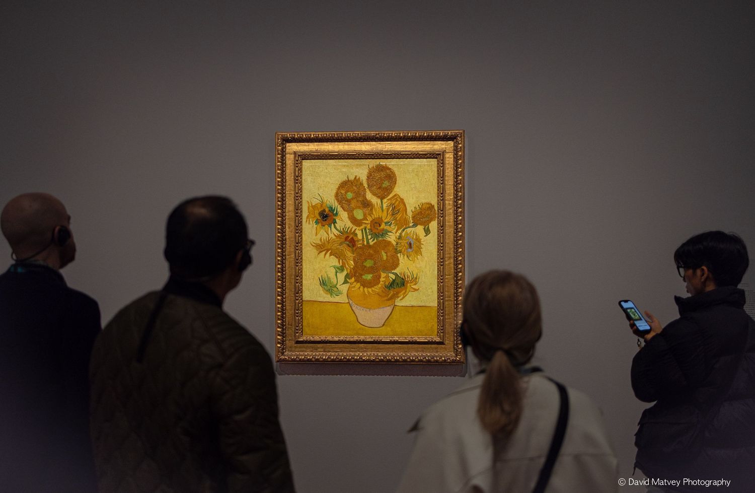 Museum visitors admiring Van Gogh's Sunflowers painting displayed on a gray gallery wall.