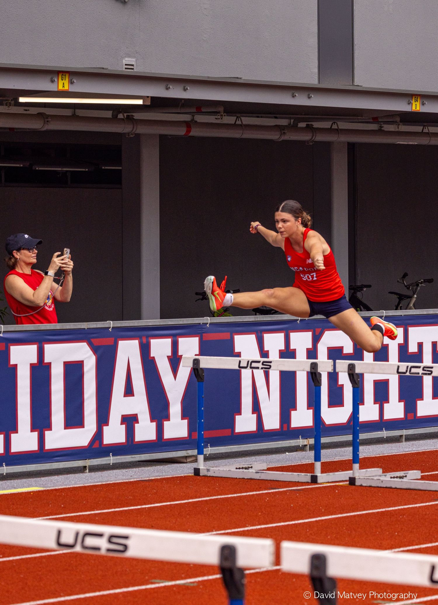 Female athlete clears hurdle during indoor track meet with Holiday Night signage visible.