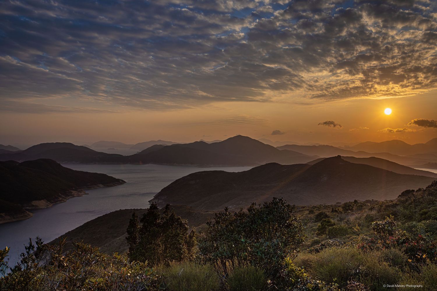 Golden sunset over misty mountain layers with a winding river valley below and dramatic cloudy sky.
