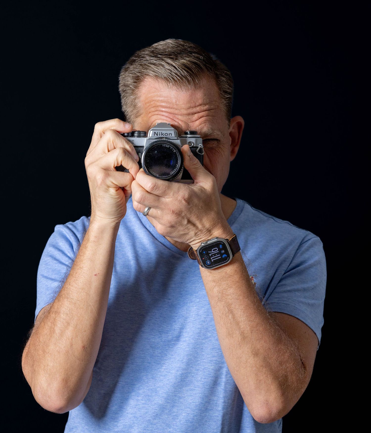 Man in blue t-shirt holding vintage film camera up to his face against dark studio background.